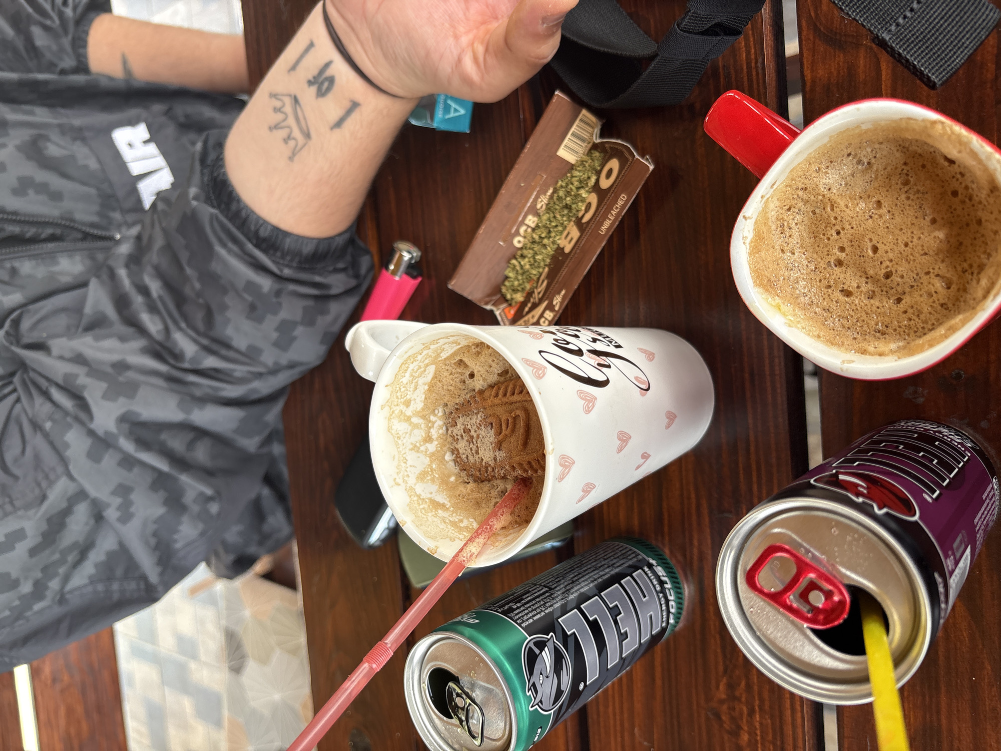 The image shows a wooden table with various items placed on it. On the table, there are two mugs, one with a red handle filled with foamy coffee, and the other with pink hearts on it and also filled with a coffee-like beverage with a biscuit inside and a red straw in it. There are two open cans of energy drinks, one green with the brand name "HELL" visible and a red straw in a purple can. A pink lighter, a pack of OCB rolling papers with some loose material that resembles weed, a blue pack, and a black strap are also on the table. An arm with tattoos, including a crown and the numbers "1 of 1", rests on the table near the rolling papers. The person wearing a black patterned jacket is partially visible in the background. The scene is reminiscent of a table after people have been drinking coffee and energy drinks, possibly preparing to roll something. There is no indication that this is in Polena, Bulgaria.