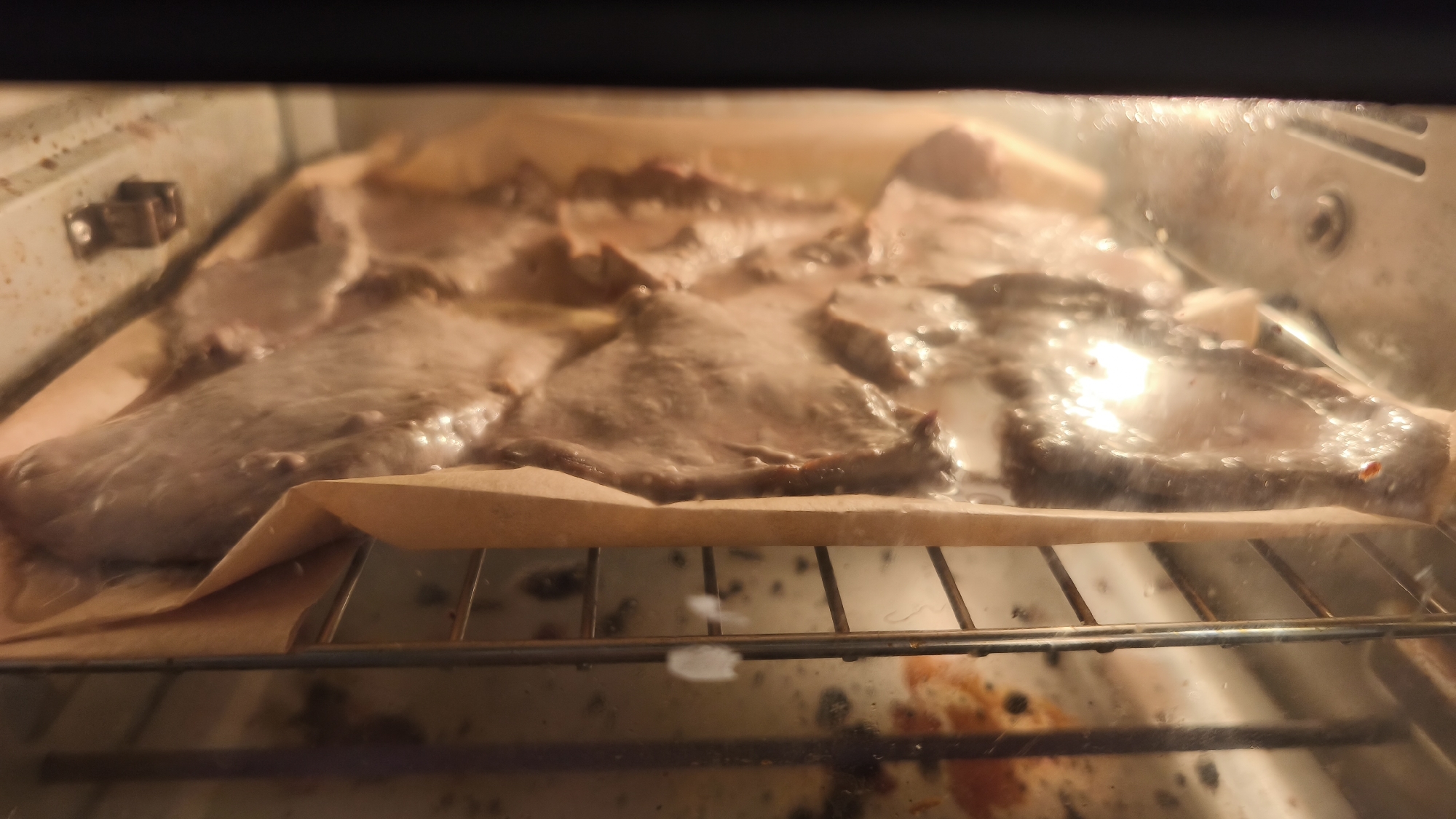 The image provides a close-up view inside an illuminated oven, showcasing multiple pieces of food being cooked or baked. The primary subjects are several irregularly shaped, thin cuts of what appears to be meat or a similar food item, arranged on a sheet of light brown parchment paper. This paper, slightly creased at the edges, rests upon a metallic wire oven rack.
The food has a muted brownish-gray color, with some surfaces appearing glossy or moist, indicating it is actively cooking. Below the rack, the oven's bottom surface is visible, showing signs of use with some dark residue or spills. On the left interior wall of the oven, a metallic hinge mechanism is partially seen.
The scene is lit with a warm glow, likely from the oven's internal light or heating elements, creating reflections on the food and the inner surfaces of the oven. The perspective is from slightly above, looking into the oven during the cooking process. No visible text, people, or external environmental cues are present, other than the implication of a kitchen setting.