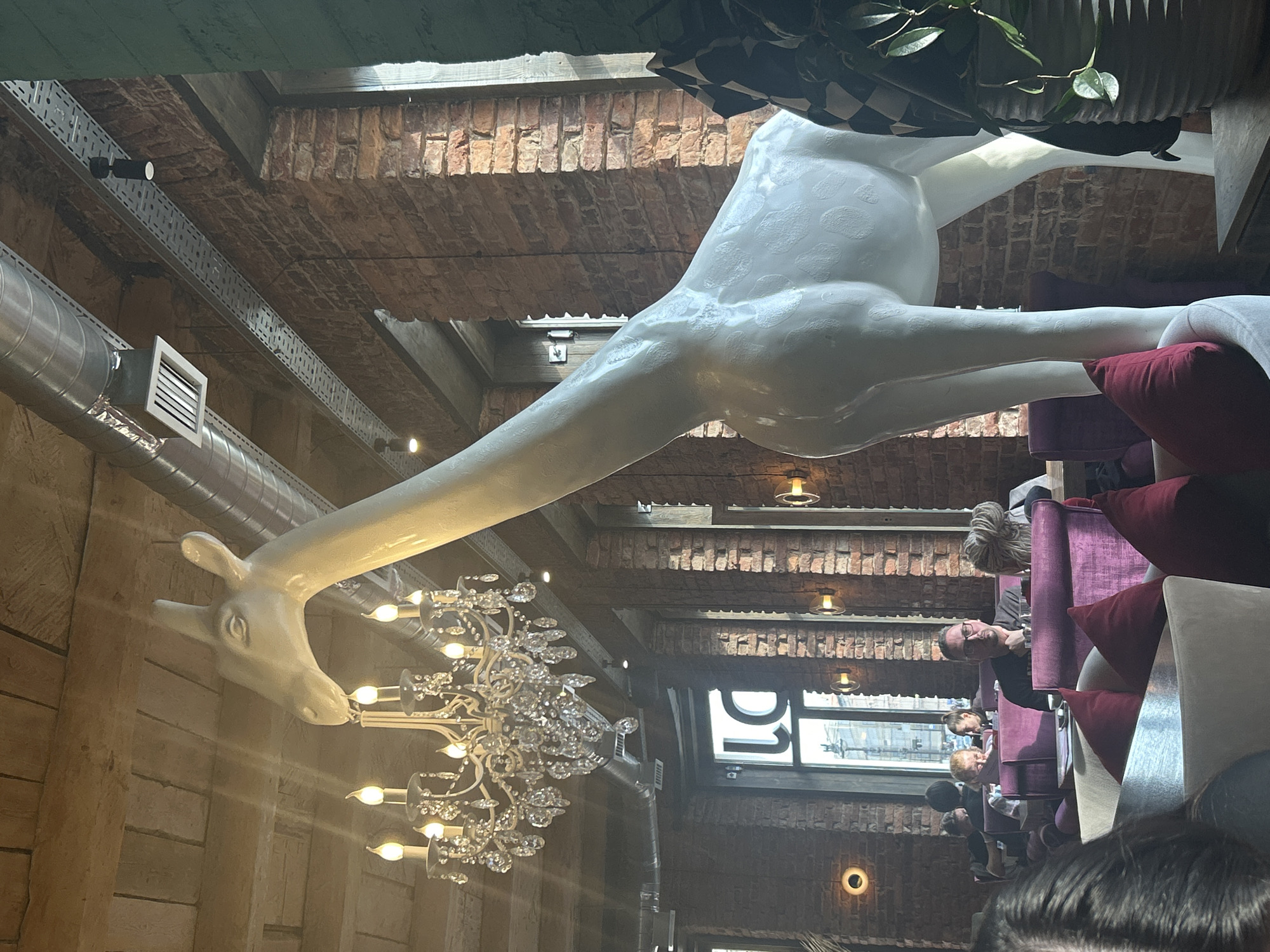 In an indoor setting, a large, white giraffe statue stands prominently in the foreground, facing right. The statue's textured surface mimics the spots of a real giraffe. Behind the statue, a brick wall forms the backdrop, with several windows letting in natural light. Exposed ventilation ducts and industrial-style light fixtures are visible along the ceiling and upper wall. In the middle ground, diners are seated at tables, with some engaged in conversation. A crystal chandelier hangs from the ceiling, adding a touch of elegance to the otherwise industrial-chic environment. The overall scene suggests a restaurant or cafe with a unique decorative theme.