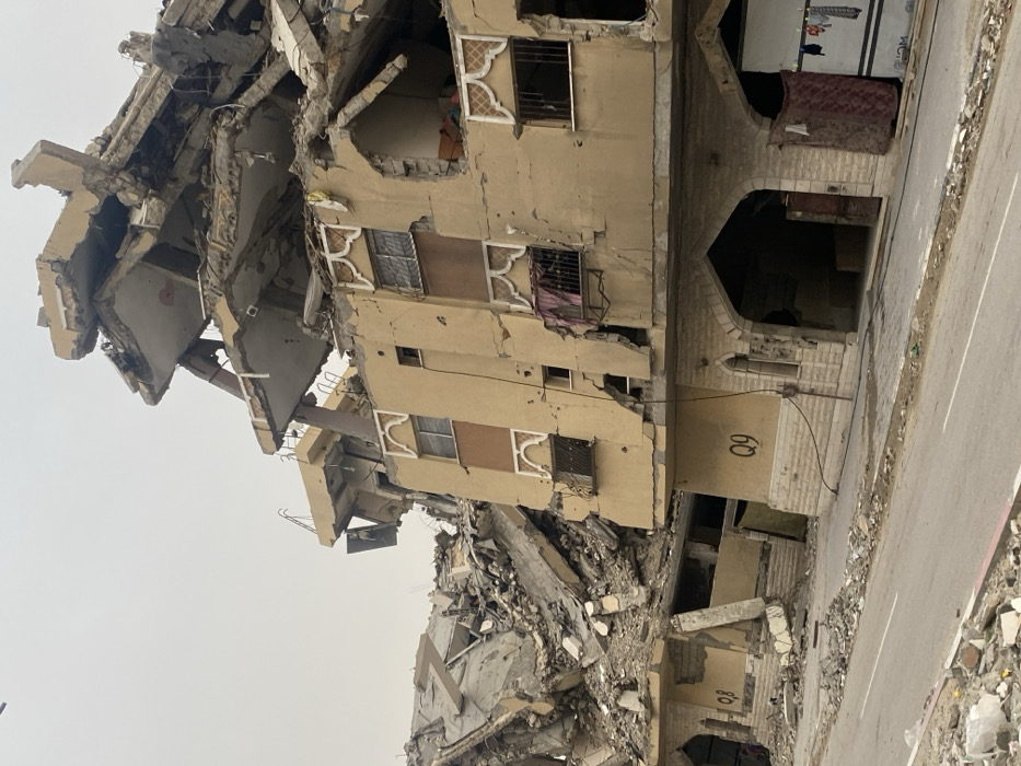 The image depicts a scene of widespread devastation, showcasing the aftermath of a destructive event. The primary focus is on a multi-story building, rendered in a light beige or sandy color, which stands partially upright but is severely damaged.
The central building exhibits significant structural collapse, particularly on its upper left side, where floors and walls have crumbled inwards and outwards, exposing twisted rebar, broken concrete slabs, and remnants of interior spaces. Many windows on the building are shattered, boarded up, or have their frames bent and broken. Some windows feature decorative, intricate white grates, adding a touch of traditional architectural style. Cracks spiderweb across the facade of the building, indicating structural stress. On the ground floor, a distinctive arched entranceway, built with what appears to be light-colored stone or brick, leads into a dark interior. A black cable runs vertically along the right side of this section of the building. Clothes or fabric items are visible hanging from a broken window or balcony railing on one of the upper floors, and what looks like a mattress is propped near the arched entrance, suggesting recent occupancy or attempts to salvage belongings.
To the left and foreground of the central building, a massive pile of rubble extends, consisting of concrete debris, broken masonry, and bent metal, indicating the complete collapse of adjacent structures. This area is a chaotic mix of shattered building materials.
A paved road runs along the right side of the scene, with a white line partially visible on its surface. The edge of the road is strewn with smaller pieces of debris, concrete fragments, and dirt, suggesting the impact extended beyond the immediate structures.
The sky overhead is overcast and light grey, providing a flat and somber lighting to the scene. There are no people visible in the image, conveying a sense of abandonment and stillness after the catastrophe.
Visible text on the facade of the central building includes "Q9" prominently displayed in black lettering near the arched entrance. Further down and partially obscured by rubble, the text "Q8" is also faintly visible.
The overall impression is one of profound destruction and tragedy, highlighting the severe damage inflicted upon residential or commercial buildings.