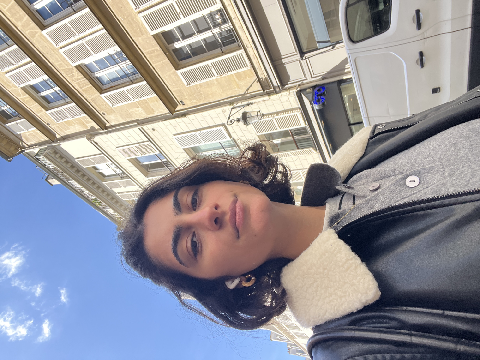 A young woman is taking a selfie on a sunny day. The background includes a building with multiple windows and shutters, typical of Parisian architecture. A white van is partially visible on the right. It appears to be in an urban setting in Paris. She is wearing a leather jacket with a faux-fur collar and has curly dark hair, and an earbud in her right ear.