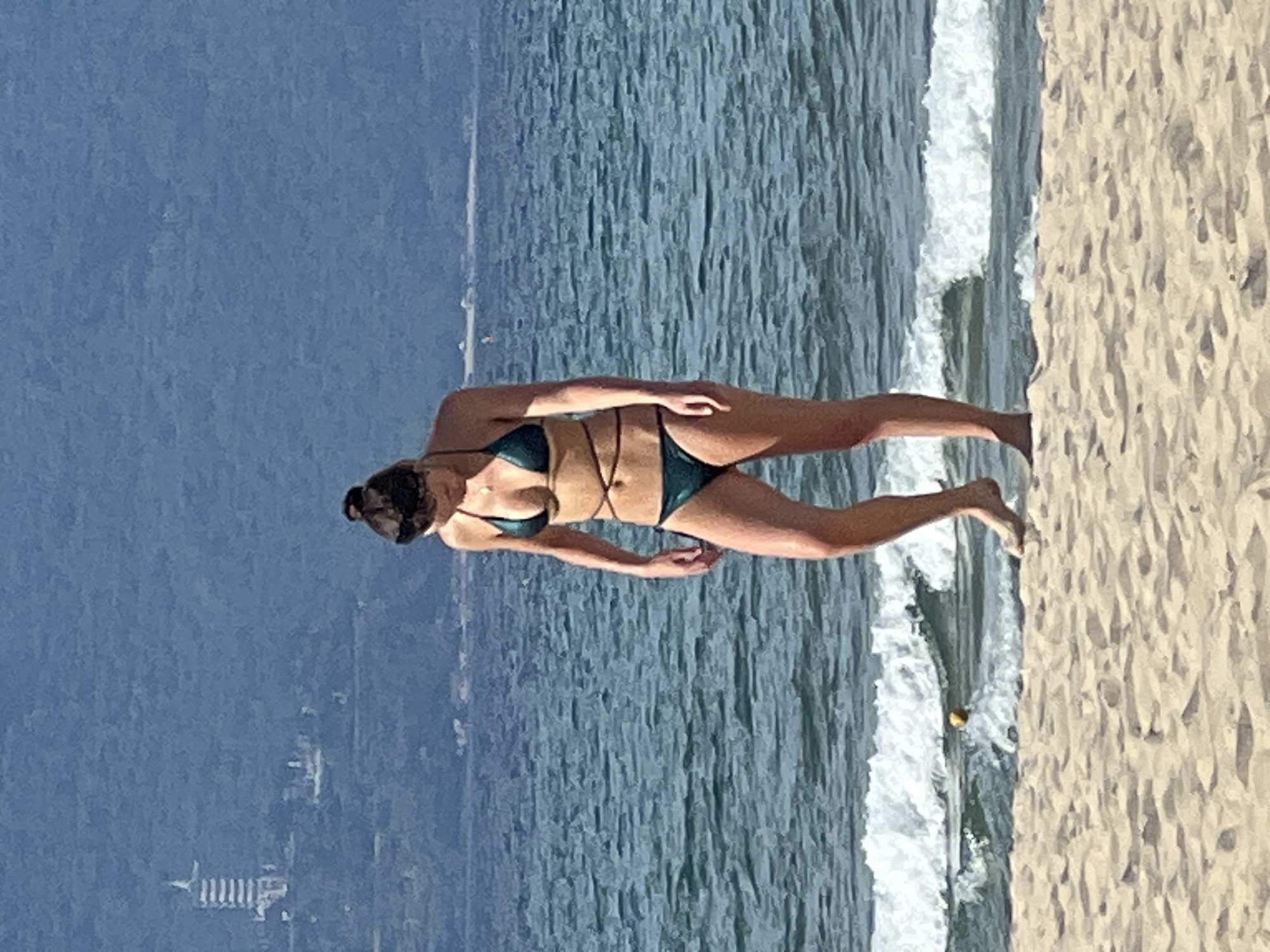 A woman in a dark green bikini stands on a sandy beach, facing the ocean. The waves are breaking gently on the shore. In the background, a dense, green hillside is visible, with a white, multi-tiered structure, resembling a pagoda or tower, situated near the top of the hill. The sky is a hazy blue. The scene is likely Da Nang, Vietnam, given the typical mountainous coastal landscape and the architectural style of the building.