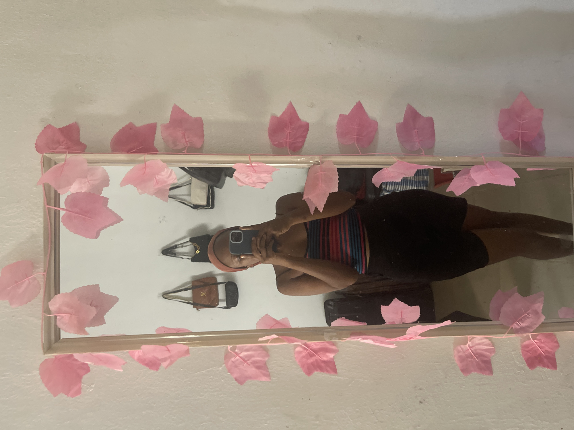 The image shows a young woman standing in front of a full-length mirror, taking a selfie. She is wearing a striped tank top and dark shorts, and a rust-colored headwrap. The mirror is decorated with a string of artificial pink leaves, which are also attached to the wall. In the reflection of the mirror, several handbags are visible hanging on the wall behind the woman. The room appears to be a bedroom, with a white wall and a wooden frame around the mirror. The lighting suggests it is daytime. The setting is likely a private residence in Jos, Nigeria, given the user's prompt.