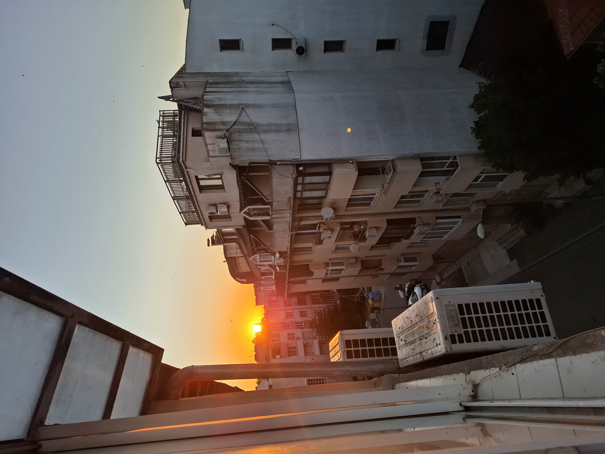 An outdoor, eye-level, vertical, medium shot captures a sunset viewed from a balcony in the city of Burgas, Bulgaria. The sun, a bright orange orb, is low on the horizon, casting a warm glow over the urban landscape. Apartment buildings of varying heights and architectural styles dominate the scene, their facades detailed with windows, balconies, and air conditioning units. In the foreground, a white, rectangular air conditioning unit sits on a concrete ledge, partially illuminated by the setting sun. A metallic awning with translucent panels extends from the left side of the frame, creating a frame for the view. The sky transitions from a pale orange near the horizon to a soft blue overhead, with a few small birds visible in flight. A street is visible between the buildings, with parked cars and a street lamp. The overall impression is one of a quiet evening in a densely populated urban area.