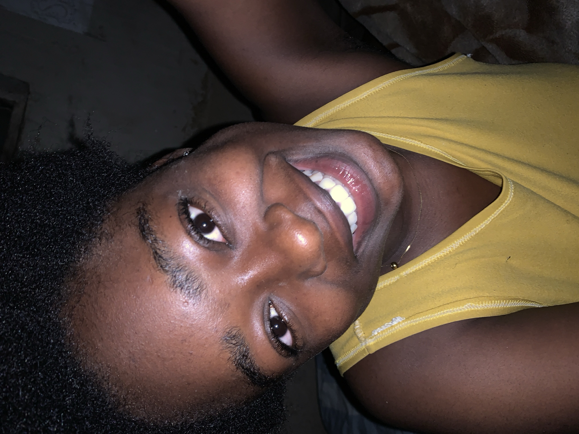 A close-up, overhead shot shows a young Black person with dark skin, black curly hair, and a bright yellow tank top. The person is smiling with their mouth open, revealing white teeth. They are looking directly at the camera. The background is dark and out of focus, suggesting an indoor setting, possibly a bedroom in Rafin Par, Nigeria. The lighting is dim, casting shadows on the person's face and the surrounding area.