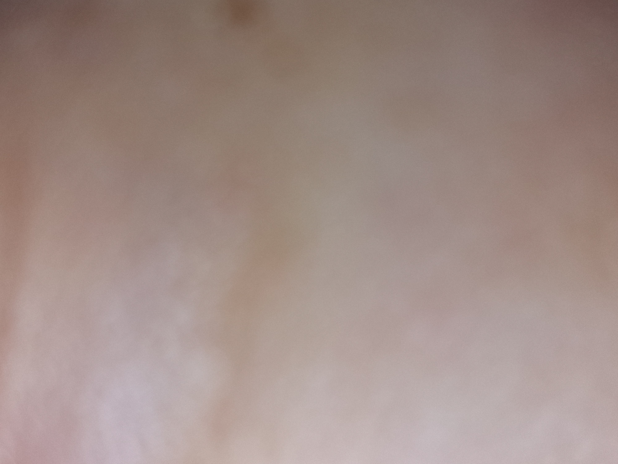 The image is extremely blurry and lacks any distinguishable features. It appears to be an out-of-focus shot of a light beige or skin-toned surface with subtle variations in color and texture. There are no discernible people, objects, or environmental details visible. The location is indeterminable.