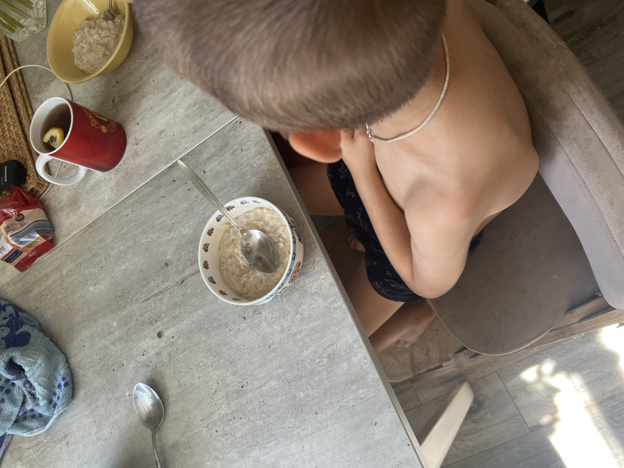 A top-down medium shot captures a young boy from the shoulders up, seated at a gray, textured table. He is wearing black shorts and a silver chain around his neck. His back is to the camera, and his head is turned away. On the table, there are two bowls of oatmeal, one with a spoon in it, a red mug with tea and a lemon slice, and a blue and white striped towel. A portion of a red and white package is visible in the background. The setting appears to be a domestic environment, likely a kitchen or dining area. No specific details about Volgograd, Russia, are discernible from the image itself.