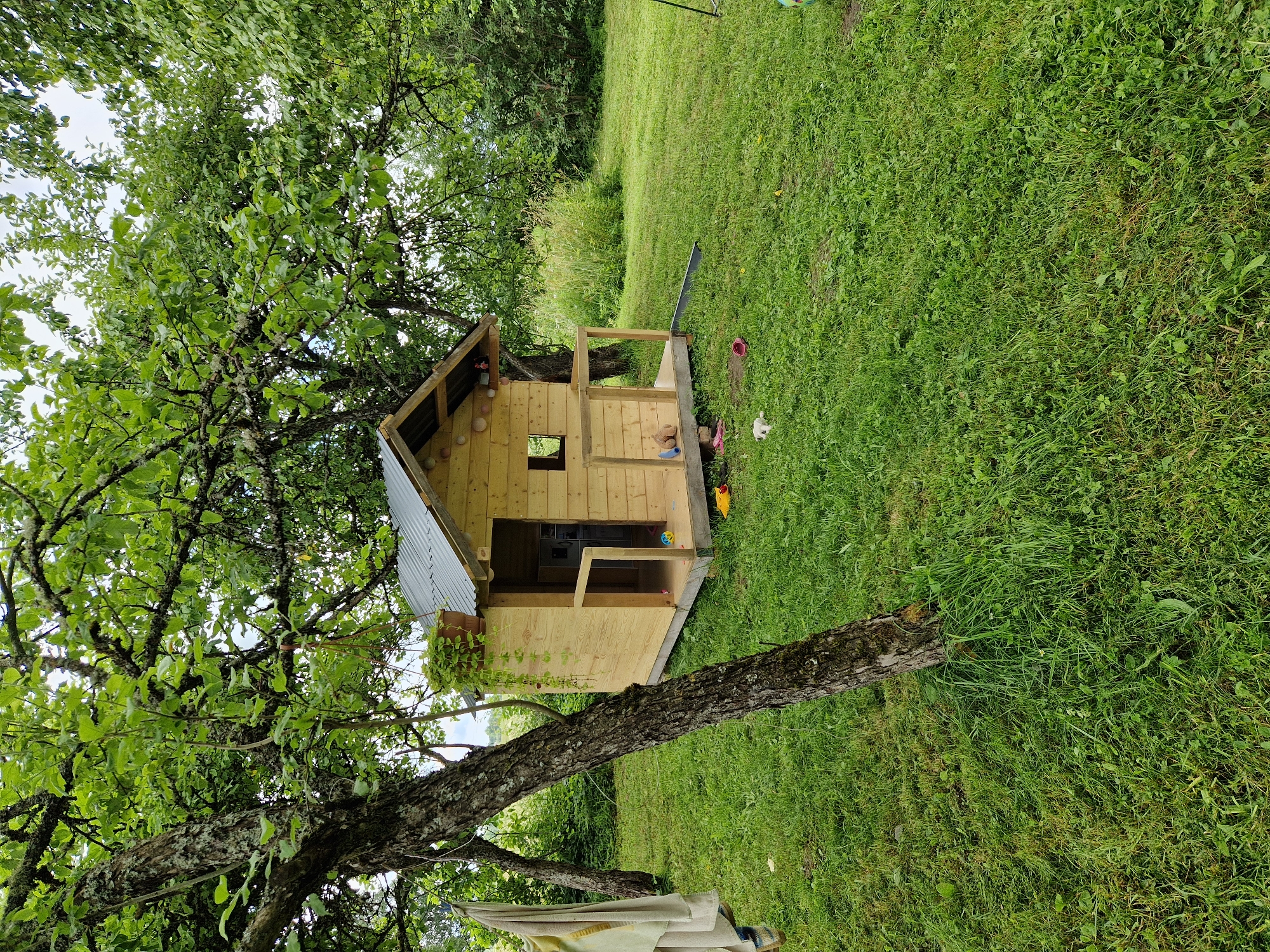 An outdoor, eye-level, medium shot captures a wooden playhouse situated on a grassy lawn beneath the canopy of a mature tree. The playhouse, made of light-colored wood, features a corrugated metal roof, a small window, and an open doorway leading to a porch with a railing. Toys, including a yellow duck, a white stuffed animal, and a pair of pink shoes, are scattered on the grass in front of the playhouse. A beige towel hangs from the trunk of the tree on the left side of the frame. The environment appears to be a rural or suburban backyard, likely in the city of Beja, Latvia, given the natural setting with abundant greenery and trees. The lighting suggests it is daytime.