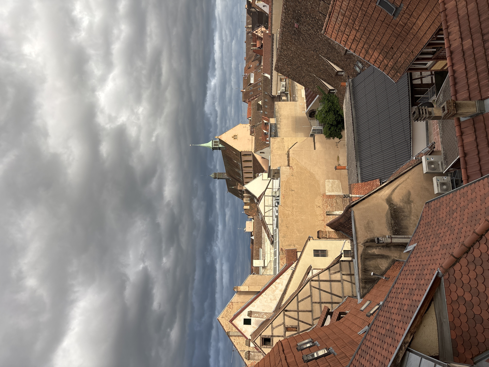 The image shows a view of rooftops in the historic district of Colmar, France, under a dramatic, overcast sky. The architecture features a mix of traditional Alsatian half-timbered buildings with exposed wooden beams and lighter-colored facades, alongside buildings with terracotta tiled roofs and plastered walls. A prominent church with a green steeple and a bell tower is visible in the background, its ochre-colored walls catching the light. The foreground is dominated by the red-tiled roofs of various buildings, some with visible dormer windows. The overall impression is one of dense urban development with a strong sense of history. There are no people or animals visible in the image. The environment appears to be a residential or mixed-use urban area.