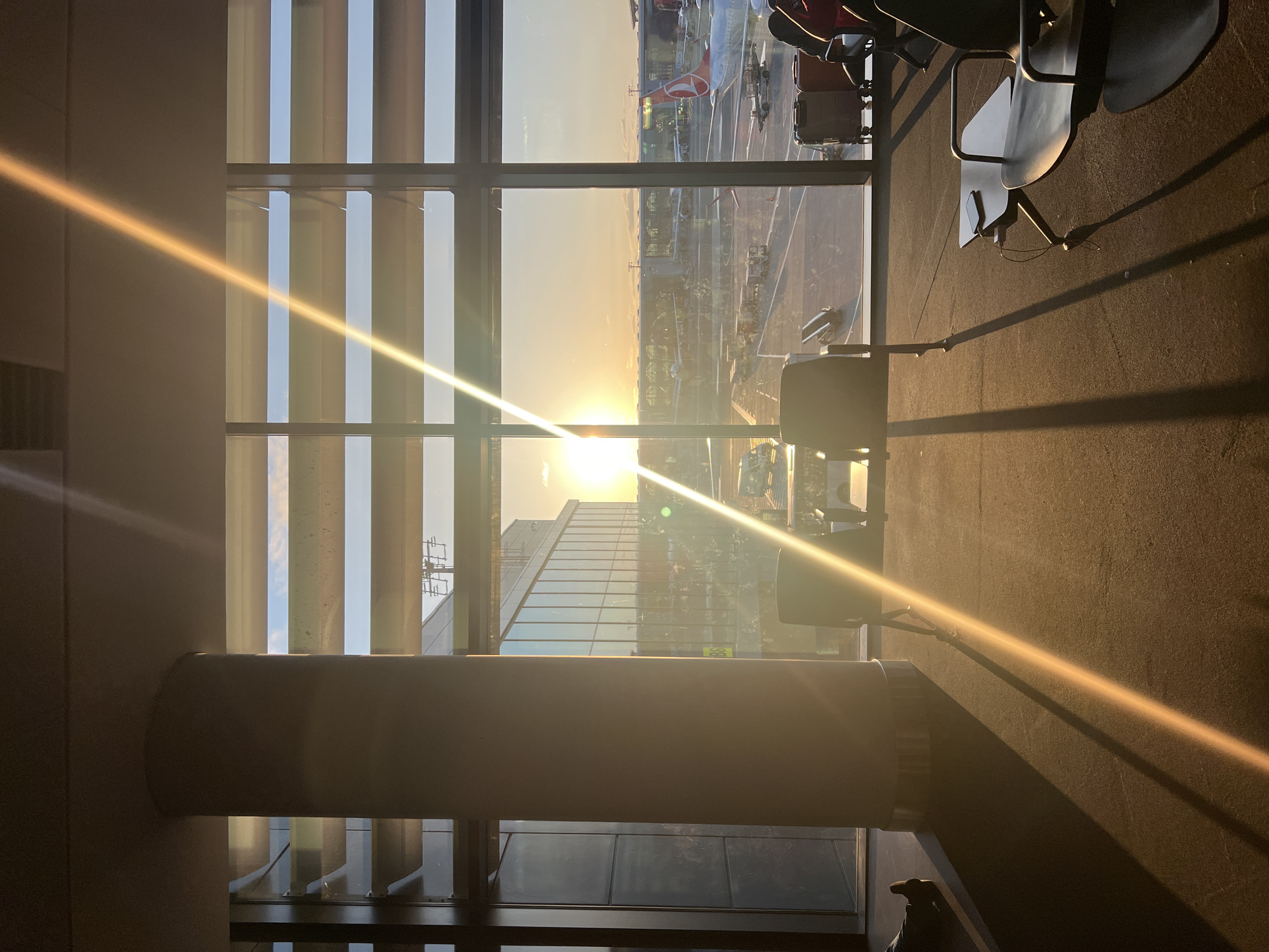 An interior view of an airport terminal in Arnavutköy, Turkey, captures a striking sunset through a large window. The sun, low on the horizon, casts a golden light that streams across the scene, creating long shadows and lens flares. Several airport chairs are visible, some occupied by people, while others are empty. Luggage is present near some of the seats. Outside the window, the airport tarmac is busy with aircraft and ground crew. The architectural elements of the terminal, including a large cylindrical pillar and the reflective glass facade of the building, frame the view of the exterior.