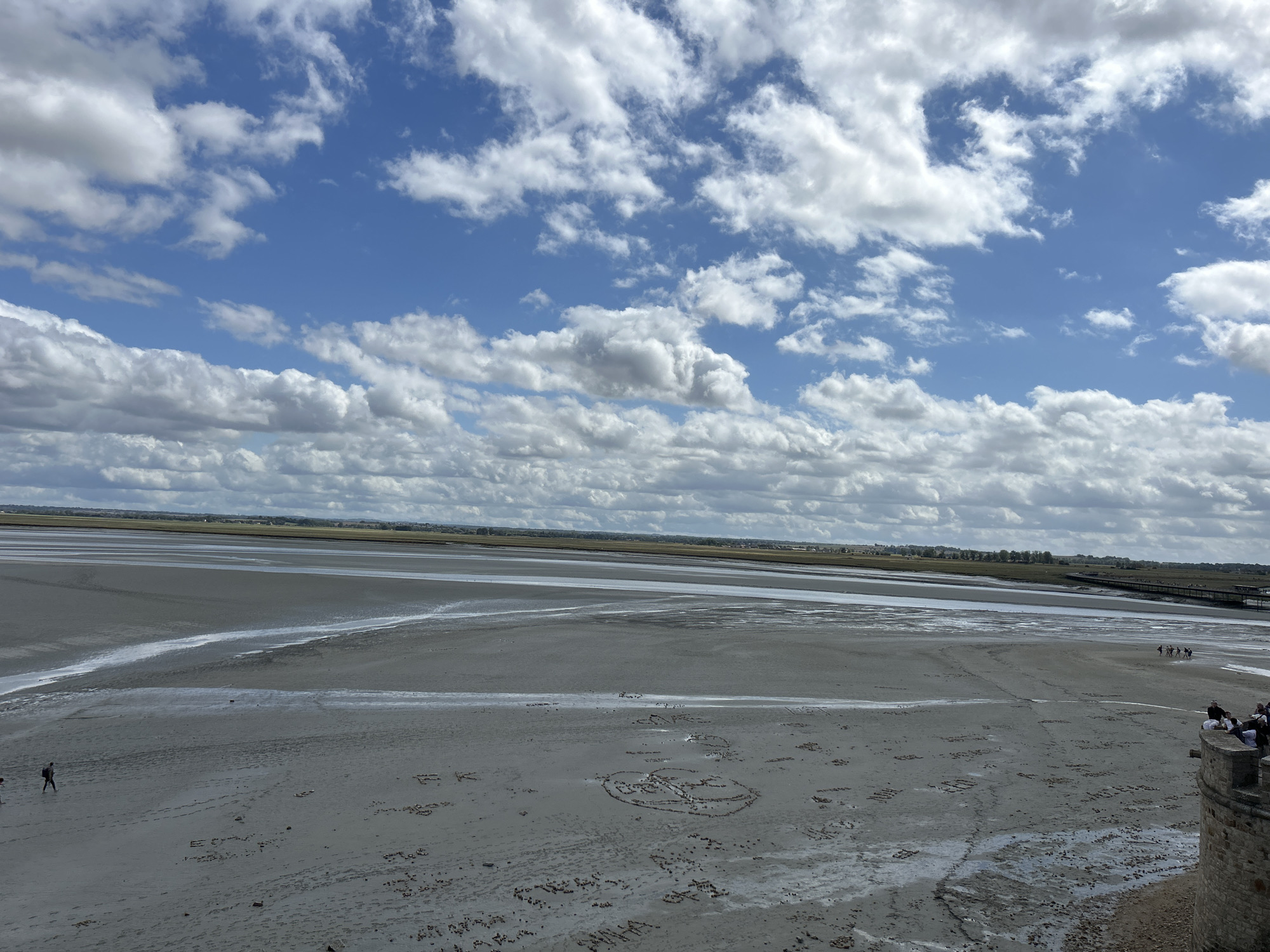 An aerial long shot captures a vast expanse of wet sand under a partly cloudy sky. The sand, which appears to be a mudflat, is etched with various words and designs. In the distance, a strip of land with vegetation and buildings stretches horizontally, bordered by a body of water. A wooden walkway or bridge extends into the water on the right side of the frame. Several small figures are scattered across the sand. On the far left, a lone individual walks away from the viewer. Closer to the center, a group of people are walking towards the right. On the right side of the image, a stone structure, possibly a battlement or tower, juts into the frame from the bottom right corner, with several people standing on top of it. The sky is a vibrant blue, filled with fluffy white clouds, some of which are darker and appear heavier. The overall impression is a wide, open landscape at low tide.