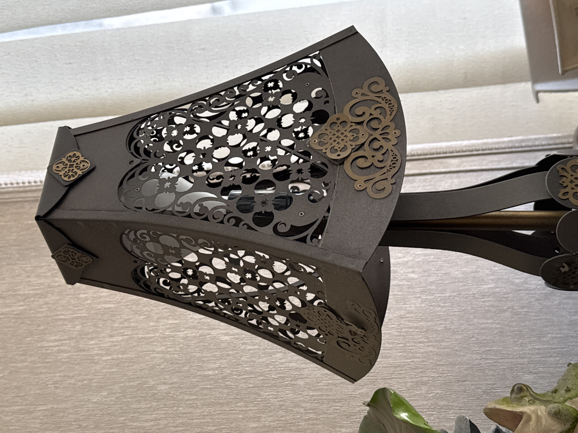 The image shows a close-up of a dark gray, geometrically shaped lampshade with intricate cut-out patterns. The lampshade has decorative gold embellishments at the top corners and along the bottom edge. The lampshade is attached to a dark stand with a visible central metal rod. In the background, a textured light beige wall is visible to the left, and sheer white curtains are visible to the right, suggesting an indoor setting, possibly a home or office. A portion of a green leafy plant and a decorative frog figurine are visible in the lower left corner. The lighting suggests a natural light source from the right. There are no people or animals other than the figurine in the scene. The location is not specified in the image itself and cannot be determined.