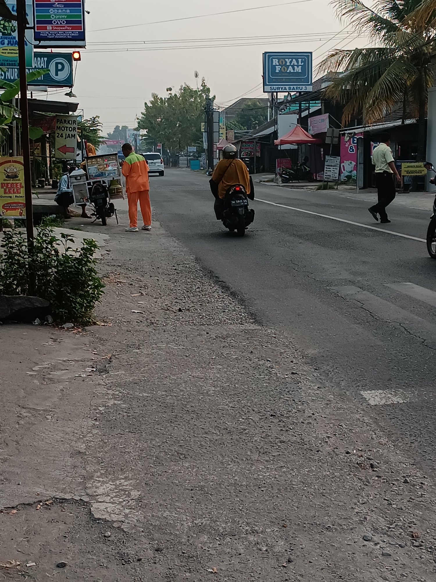 A street scene in Jetis, Indonesia, shows a man in an orange uniform standing on the side of the road next to a parked motorcycle. He is facing a food cart that is also parked on the side of the road. Behind the food cart, a person on a black motorcycle is riding away from the camera. Further down the road, another person in a light green shirt and dark pants is walking away from the camera. The road is paved with asphalt and has a white line marking the lane. On the left side of the road, there is a sidewalk with some green bushes and debris. A sign for "PARKIR MOTOR 24 JAM" (Motorcycle Parking 24 Hours) is visible on the left side of the road. Another sign, "ROYAL FOAM," is visible on the right side of the road. The sky is overcast, and the overall lighting suggests it is daytime.