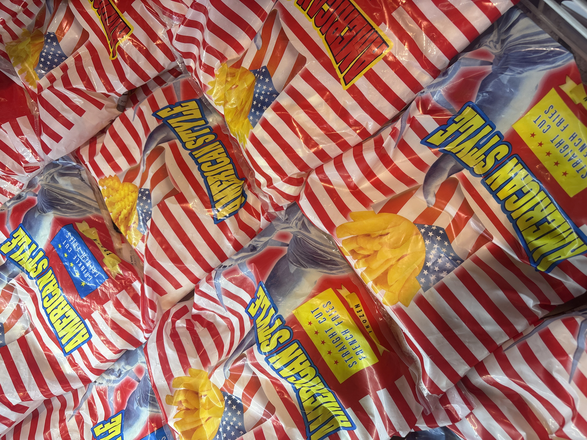 The image displays a close-up, overhead view of numerous bags of frozen french fries. The bags are predominantly adorned with the American flag design, featuring red and white stripes and a section of stars and blue. The text "AMERICAN STYLE" is prominently displayed in bold yellow lettering on many of the bags. Some bags also show images of french fries and text indicating "STRAIGHT CUT FRENCH FRIES FROZEN." The packaging is plastic and reflects light, creating a glossy appearance. The arrangement of the bags suggests they are stacked or piled together, possibly in a retail or storage setting. No people or specific environmental details of Chiang Mai City Municipality, Thailand are visible in this image.