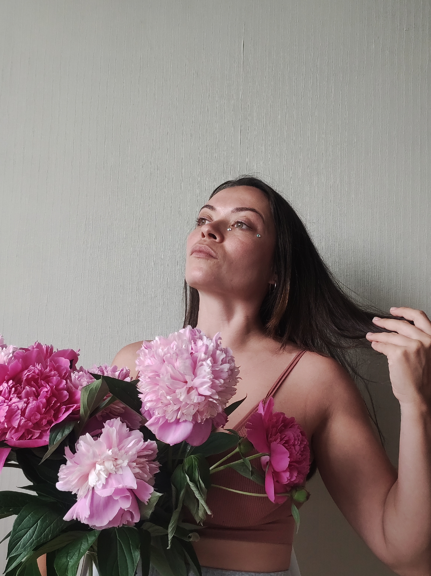 A woman is holding a bouquet of pink peonies and looking upwards. She has dark hair and is wearing a pink tank top. There are glittery stickers on her face. The background is a plain, textured wall. The image appears to be taken indoors. The specific location in Chaikovsky, Russia, cannot be determined from the image alone.