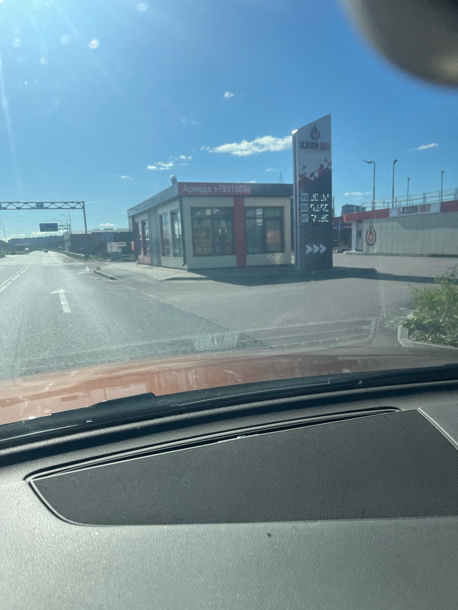 The image captures a street view of a small building, likely a convenience store or gas station, on a sunny day. The building is red and white with glass windows and doors. A sign on the building reads "Аренда +79310040", indicating a rental opportunity. To the right, a tall sign displays fuel prices, with "95" costing "53.51", "92" costing "51.71", and "ДТ" (diesel) costing "7.30". The brand name "АЗС ПЛЮС" is visible on the tall sign. The surrounding environment appears to be a road with white lane markings, a sidewalk, and some greenery. In the background, other buildings and infrastructure are visible, suggesting an urban or suburban setting. The image is taken from inside a vehicle, with the dashboard in the foreground. The sky is blue with scattered clouds.