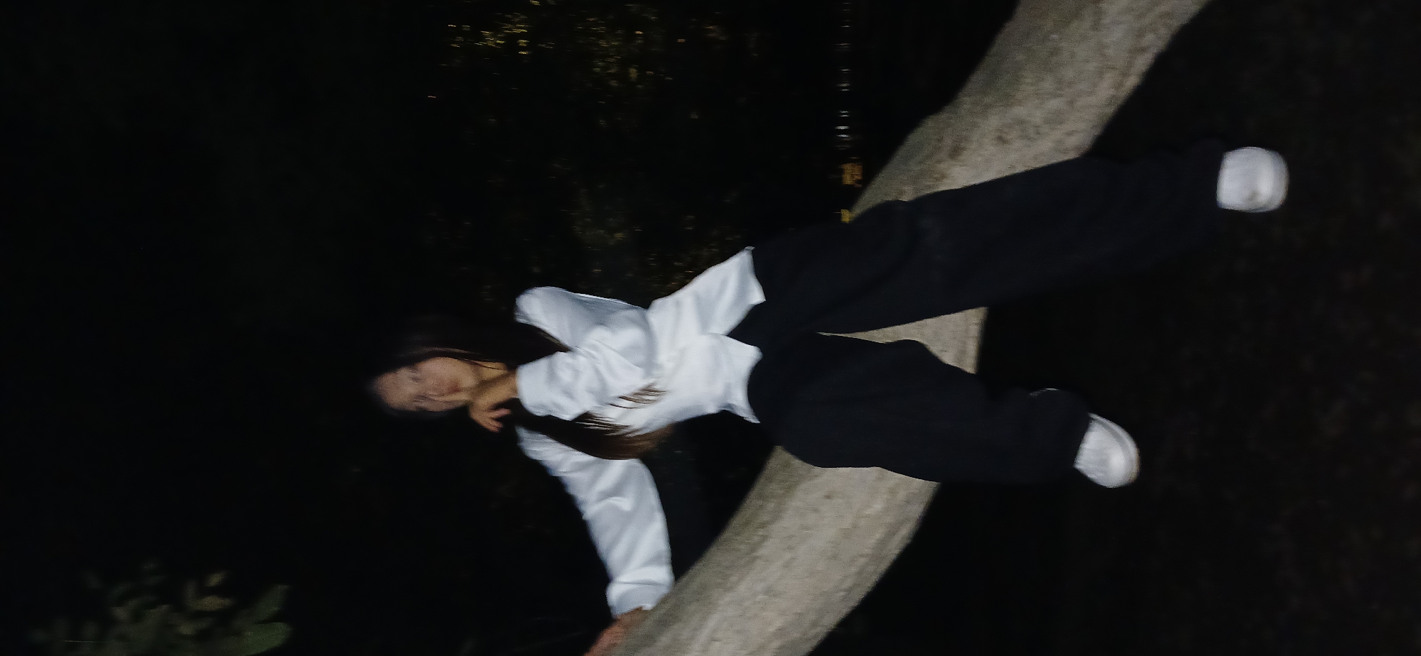 The image shows a young person with long dark hair, wearing a white collared shirt and loose black pants, sitting on a large tree branch at night. The person is positioned to the left of the frame, with their left arm resting on the branch and their right hand near their face. The background is dark and appears to be a natural environment with trees and foliage, with some distant lights suggesting a city environment. The overall image is somewhat blurry due to low light conditions and potentially motion. The specific location in Bishkek City, Kyrgyzstan cannot be determined from the visual information provided.
