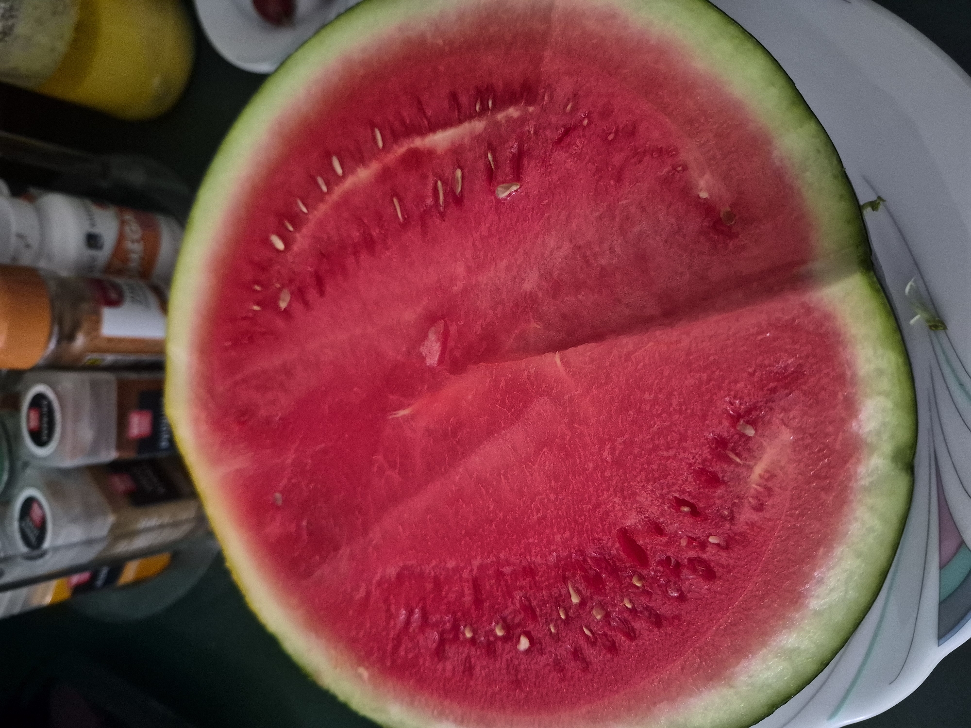 A close-up shot features a slice of watermelon placed on a white plate with a decorative rim. The watermelon is vibrant red with visible seeds and a light green rind. In the background, several spice jars and a yellow bottle are out of focus. The scene is set indoors, likely in a kitchen environment.