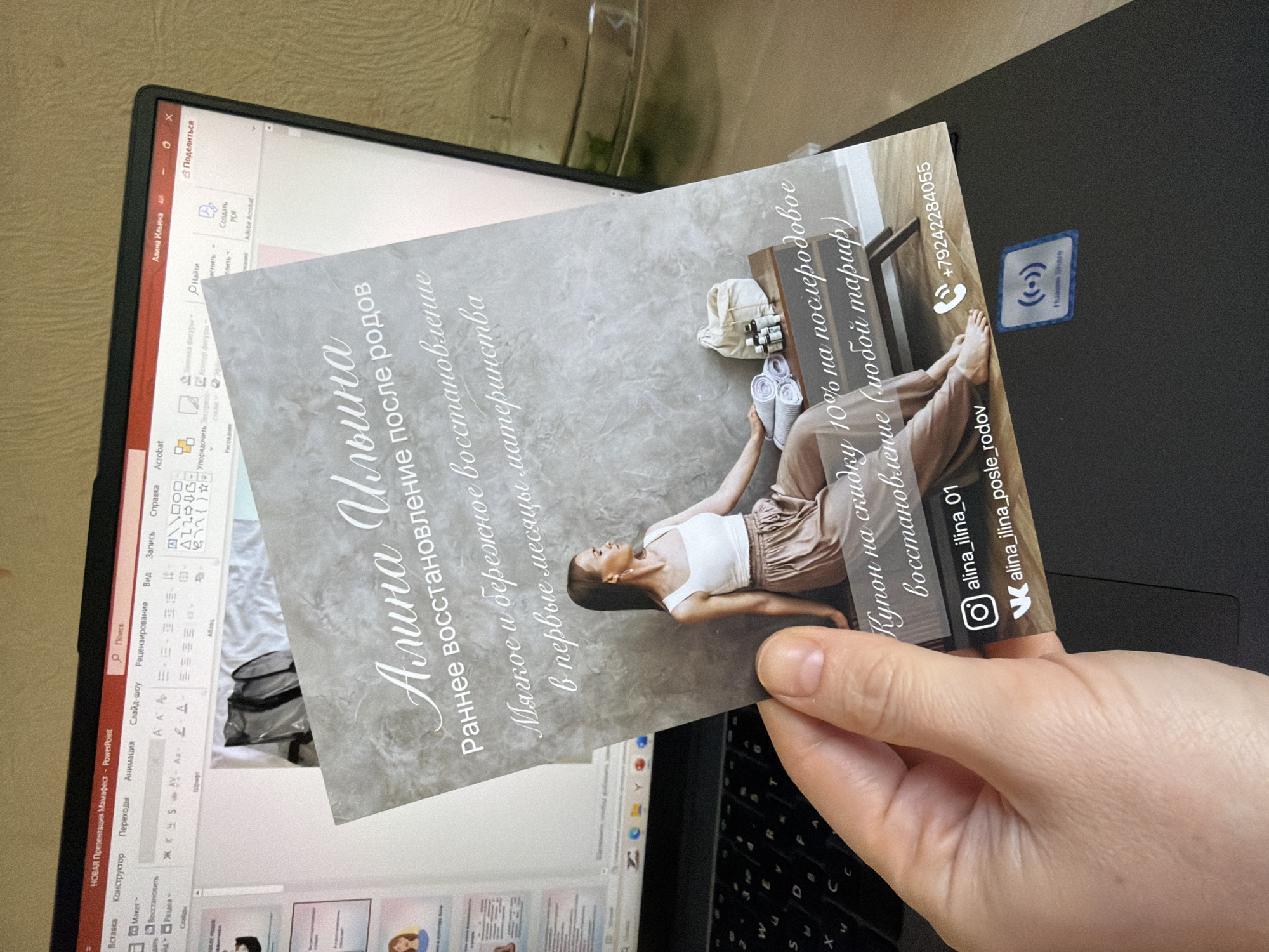 A person's hand holds a printed card in front of an open laptop screen displaying presentation software. The card features a woman sitting on a bench with towels and bottles, surrounded by text in Russian. The laptop screen shows a slide presentation interface with various text and image elements. The laptop is placed on a flat surface, likely a desk, with a blurred background that suggests an indoor environment. There is no visible indication of the city of Khabarovsk, Russia.