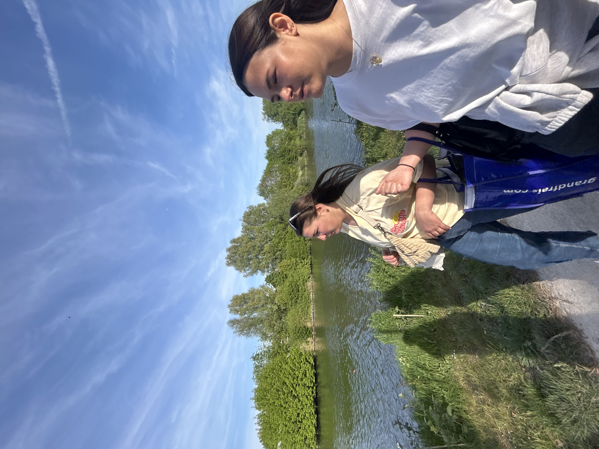 Two young women stand near the edge of a body of water, possibly a canal or river, in Marquette-lez-Lille, France. The upper woman wears a white shirt, and the lower woman wears a tan shirt and jeans. A blue bag with the text "grandfrais.com" is present. The environment includes green foliage along the water's edge and trees in the background. The sky is blue with scattered clouds.