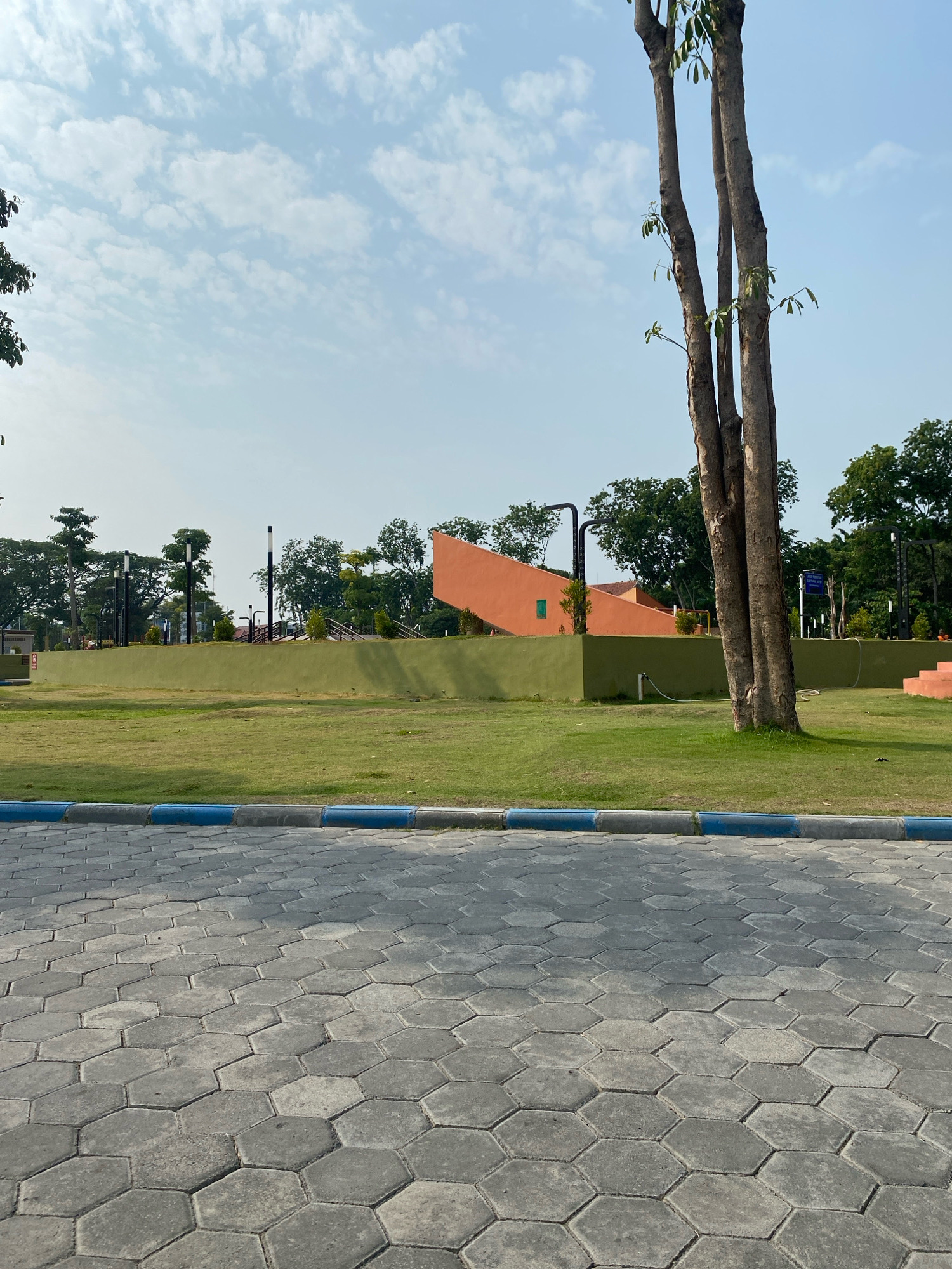 This image depicts a park area, likely located in Sidoarjo, Indonesia, given the context. The foreground is dominated by a grey hexagonal paver walkway, bordered by a curb with blue and white stripes. Beyond the walkway, a neatly manicured green lawn stretches towards a raised, green-colored embankment.
Behind the embankment, a vibrant orange structure with a modern, angular design stands out. Several streetlights with black poles and white lamps are strategically placed throughout the park. The background is filled with lush green trees and a clear sky with scattered white clouds. A tall tree with multiple slender trunks is prominently featured in the right mid-ground. The overall scene suggests a well-maintained public park designed for recreation and relaxation. There are no people visible in the image.