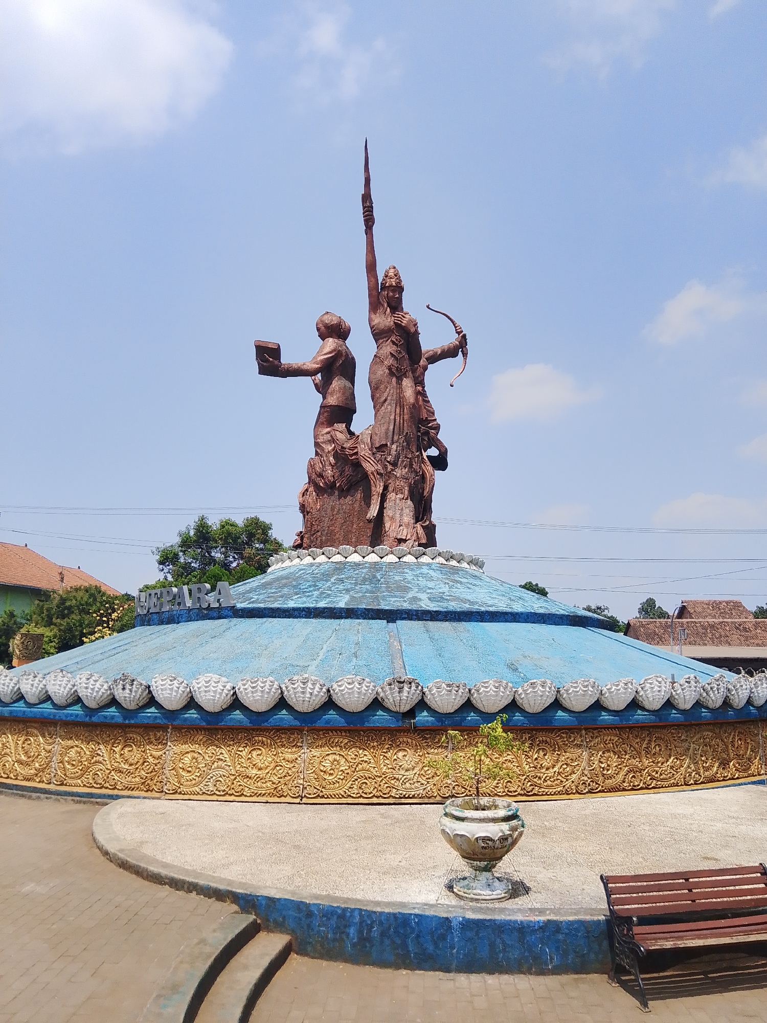 This image captures a statue in the city of Tahunan, Indonesia. The statue, made of a reddish-brown material, depicts three figures on a circular, tiered base. The central figure is a woman holding a spear aloft. To her left, another figure, possibly a woman, holds a book. To the right, a woman is drawn back, aiming a bow and arrow. The base of the statue is a vibrant blue, adorned with a decorative gold band featuring intricate patterns. Above the gold band, there is a row of white, shell-like decorations. A sign on the blue portion of the base reads "SEMARA." The ground around the statue is paved with interlocking bricks and features a concrete pathway with a curved edge and two shallow steps. A potted plant sits to the left of the pathway, and a wooden bench with black metal legs is positioned to the right. In the background, trees and buildings with tiled roofs are visible under a bright blue sky with scattered clouds.