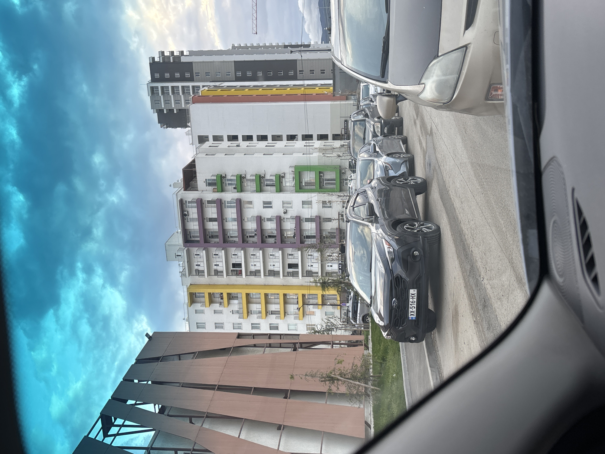 View from inside a car showing a row of parked cars on a street. In the background are several modern buildings, one with distinct horizontal color bands of yellow, purple, and green on different floors. A taller building is grey and white. A construction crane is visible above the taller building. In the foreground are the dashboard and car interior elements, partially obscuring the view. The front car has a license plate reading "XX-516-HX", which is a Georgian license plate. The location is likely in a modern residential area of Tbilisi, Georgia, given the architectural style and license plate.