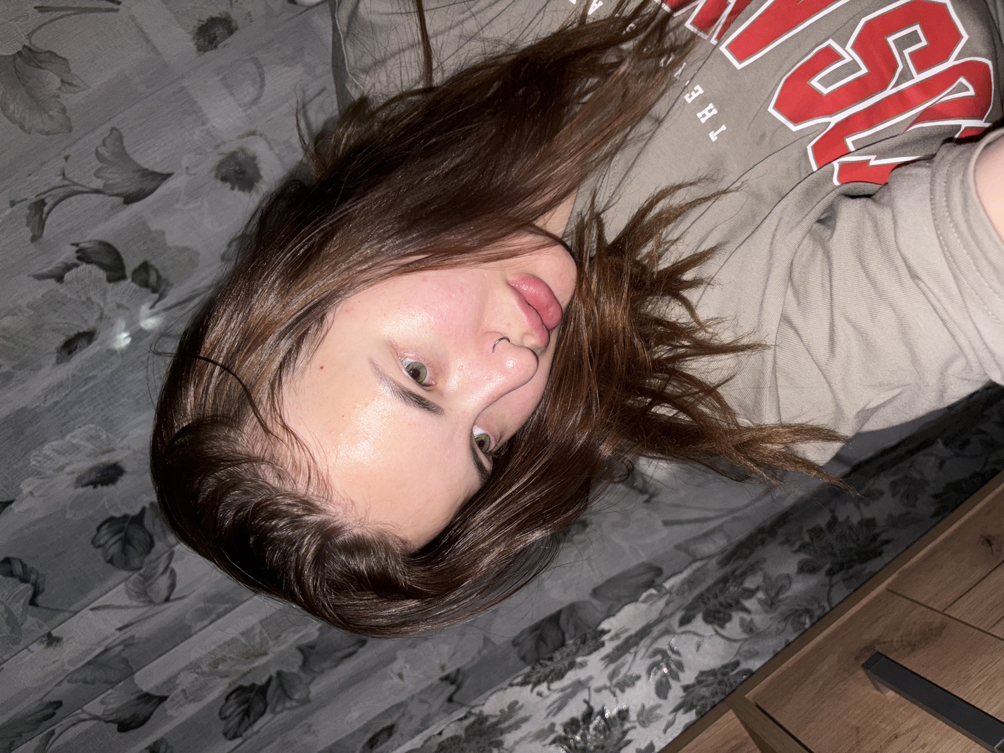 The media file captures a young individual, likely female, with long, dark brown hair spread around her head, lying on her back. The photograph is taken from an overhead perspective and is rotated, presenting her head tilted to the side. She has light-colored eyes, appearing greenish-hazel, and a small silver hoop nose ring in her left nostril. Her tongue is slightly extended, resting on her lower lip, giving her expression a casual or playful demeanor. Her skin appears fair, with some natural redness on the cheeks and forehead.
She is wearing a light gray or taupe t-shirt. Visible text on the shirt includes "THE Y" in white block letters and a large red letter "S" with a white outline, suggesting it might be part of a larger word or logo.
She is resting on bedding that features a gray background with a darker, intricate floral or leaf pattern. To the right side of the frame, a wooden surface, possibly a nightstand or other furniture, is partially visible. The lighting is soft and even, indicating an indoor setting, likely a bedroom. The overall scene conveys a relaxed atmosphere in Port-Katon, Russia.