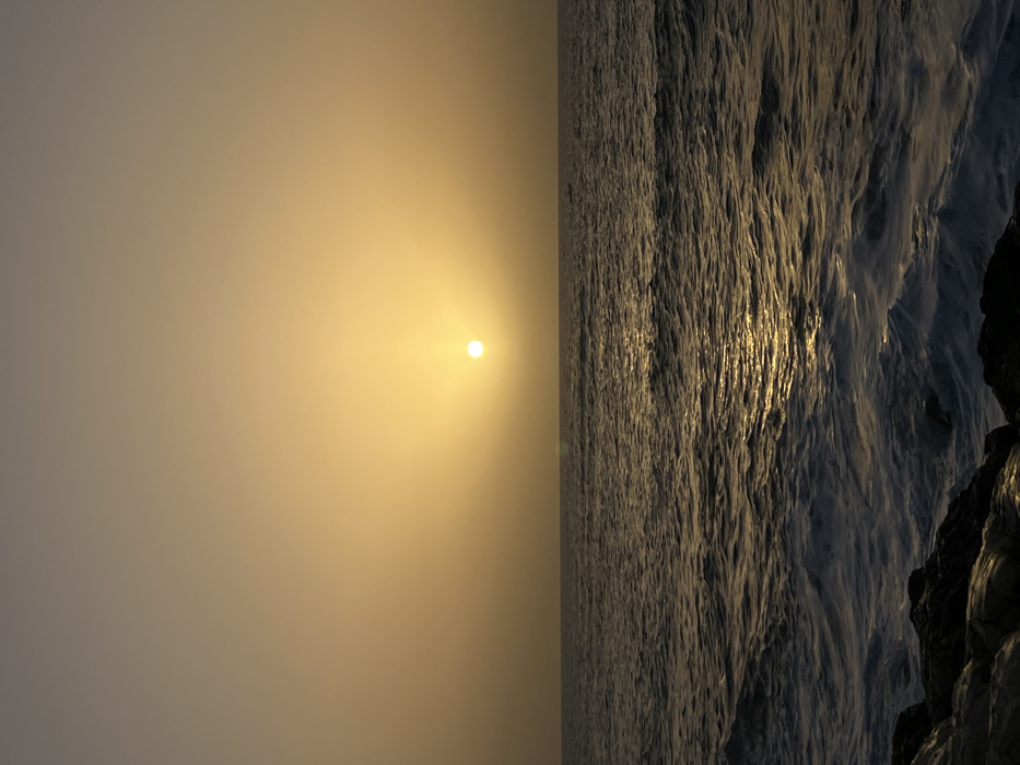 The image displays a sunset over a body of water, likely the Mediterranean Sea off the coast of Tel-Aviv, Israel. A distinct, bright, white-yellow sun orb is centered in the upper mid-right portion of a hazy, golden-orange sky, casting a broad glow. The horizon line is sharply defined, separating the sky from the ocean. The ocean surface is characterized by dark, undulating waves, with a path of shimmering golden light reflecting the sun's position. In the immediate foreground, at the bottom of the frame, dark, wet rocks are visible, with white foam from breaking waves splashing against them. No people, vessels, or artificial structures are discernable.