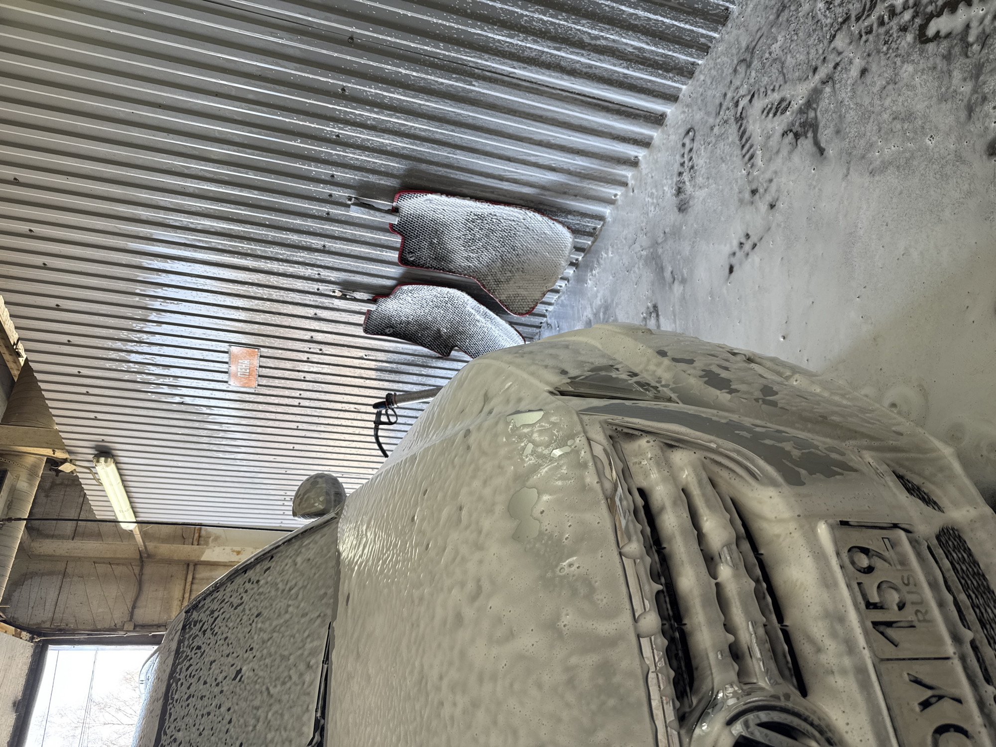 A car wash in Chaikovsky, Russia. A silver car is covered in soap suds. The license plate reads "O415XY 159 RUS". Two cleaning mitts are attached to the corrugated metal roof. Soap suds are splattered on the adjacent wall, with traces of shoe prints.