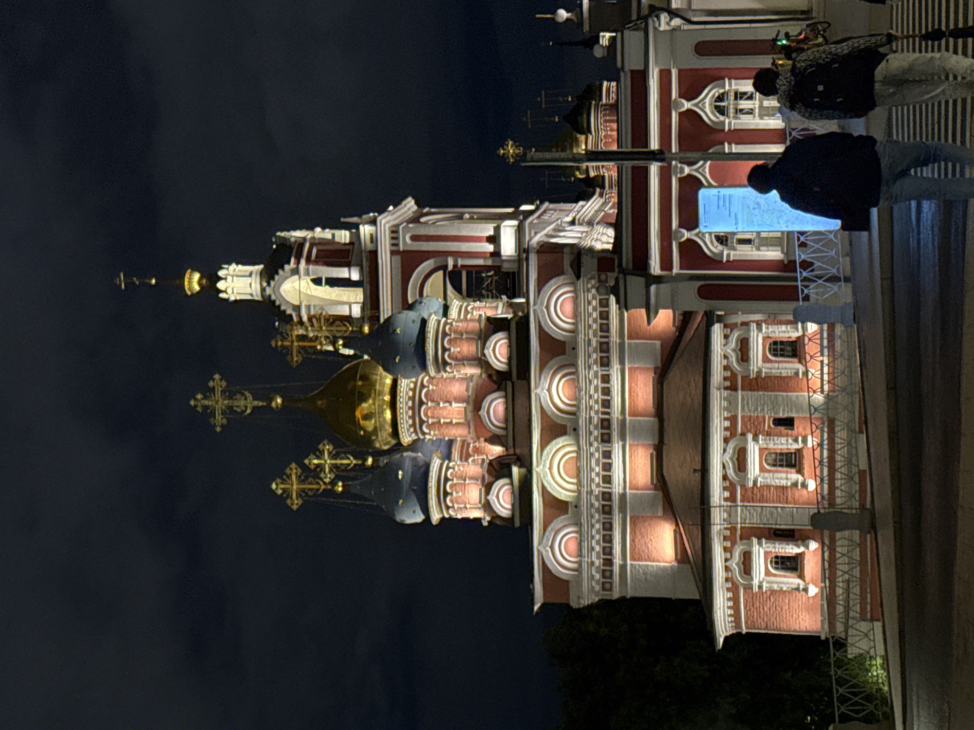 The image shows the Church of St. Nicholas at Bersenevka Embankment in Moscow, Russia, at night. The building features a red and white facade with architectural embellishments and gold and blue domes topped with Orthodox crosses. People are standing along a pedestrian walkway and looking at an illuminated information display. The background is a dark night sky.