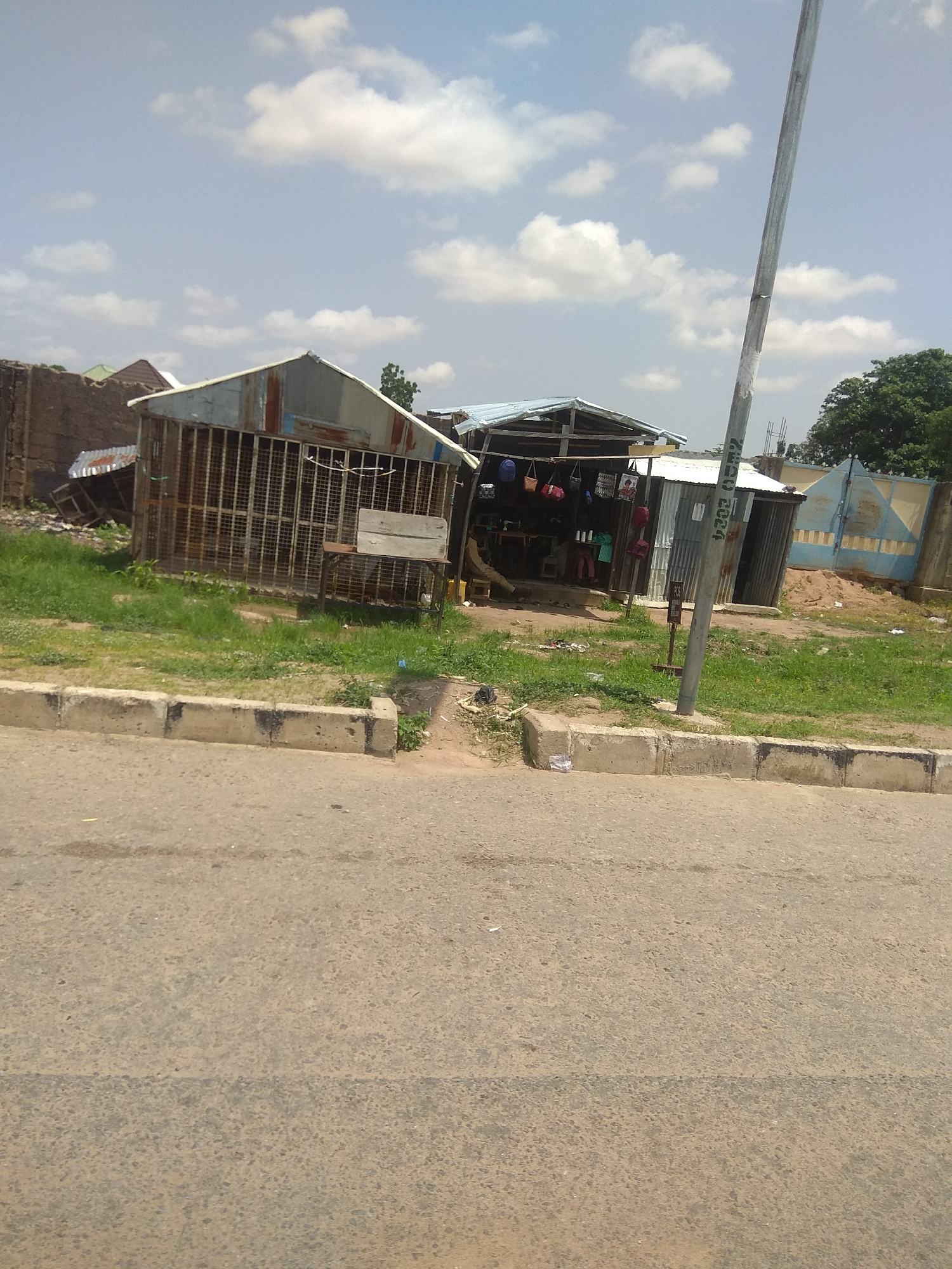 The image depicts a street scene in Jalingo, Nigeria, featuring a small roadside shop. The shop is constructed from corrugated metal and has an open front, showcasing various goods. Several bags, including backpacks and handbags, are displayed hanging from the shop's structure. A sewing machine is visible inside the shop, suggesting it might also be a tailoring establishment. A metal grate fence encloses part of the shop. In front of the shop, a paved road runs along a grassy verge, separated by a concrete curb. A tall metal pole, likely for street lighting or communication, stands near the shop. In the background, there are other structures, including a blue metal gate and trees, indicating a semi-urban or residential area. The sky is partly cloudy, suggesting daytime. There are no people visible in the image.