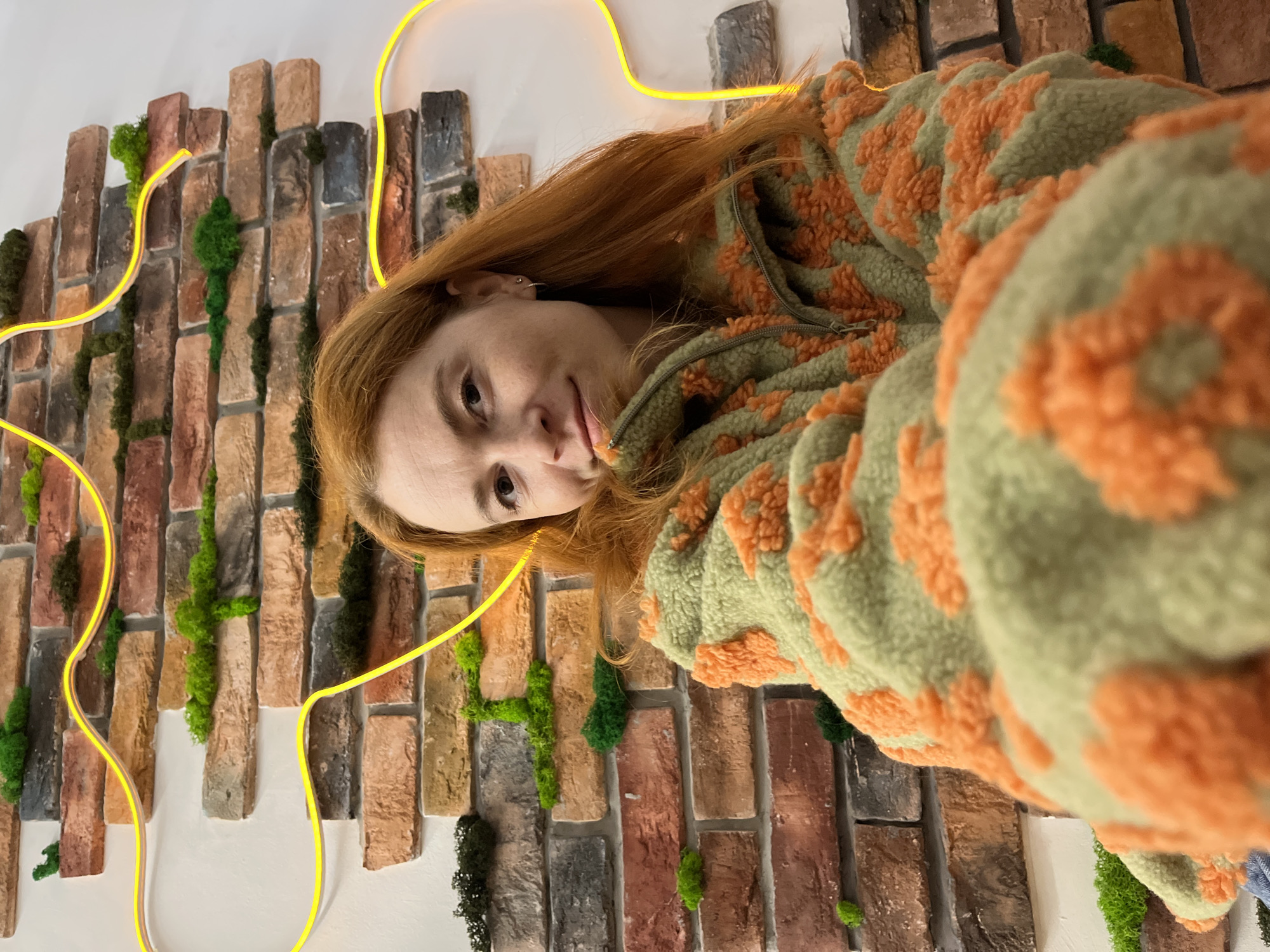 A young woman with long, auburn hair is the primary subject of this media file. She is wearing a fuzzy green jacket with an orange floral pattern. Her head is tilted upwards and slightly to the side, and she is looking directly at the camera with a gentle smile. Her gaze suggests a relaxed and possibly playful mood.
The background is a textured wall made to resemble a brick facade, with variations in brick color and size, interspersed with patches of green moss or artificial greenery. A bright yellow neon light, curved into a loose, wavy line, snakes across the frame, partially obscuring the background and framing the woman's face. The lighting appears to be artificial, given the presence of the neon light, and does not suggest a specific time of day or weather condition.
The overall scene is a close-up portrait with an artistic and somewhat quirky backdrop. The focus is on the woman's face and upper body, with the environment providing a visually interesting contrast to her. The composition, with its slight tilt and the dynamic neon line, adds a contemporary feel to the image. No text is visible in the frame. The location context of Chaikovsky, Russia, does not offer any immediate visual cues within the image itself.