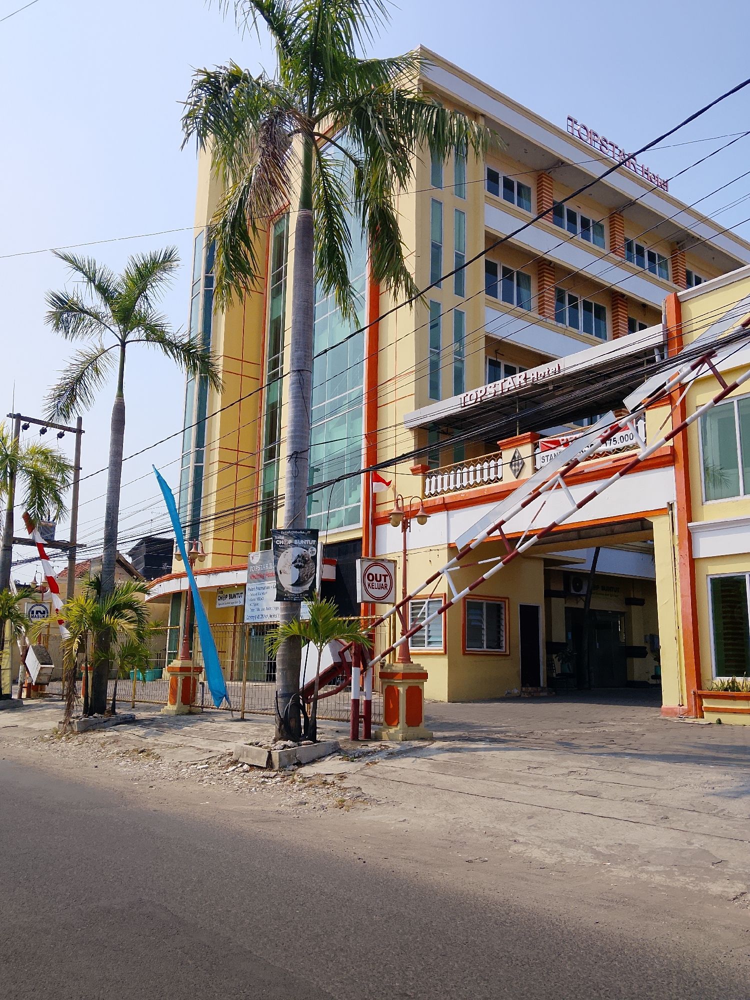 The image depicts the exterior of the Topstar Hotel in the city of Ngelo, Indonesia. The hotel is a tall, yellow, multi-story building with numerous windows. In front of the hotel is an entrance gate with a white and red striped barrier. Palm trees are visible in front of the hotel, and a street runs along the foreground. The sky is clear and blue. Signs on the building indicate the hotel's name and also display the words "MASUK" and "KELUAR," which mean "enter" and "exit" in Indonesian. A red and white flag is also visible on the left side of the image. The scene appears to be a sunny day.