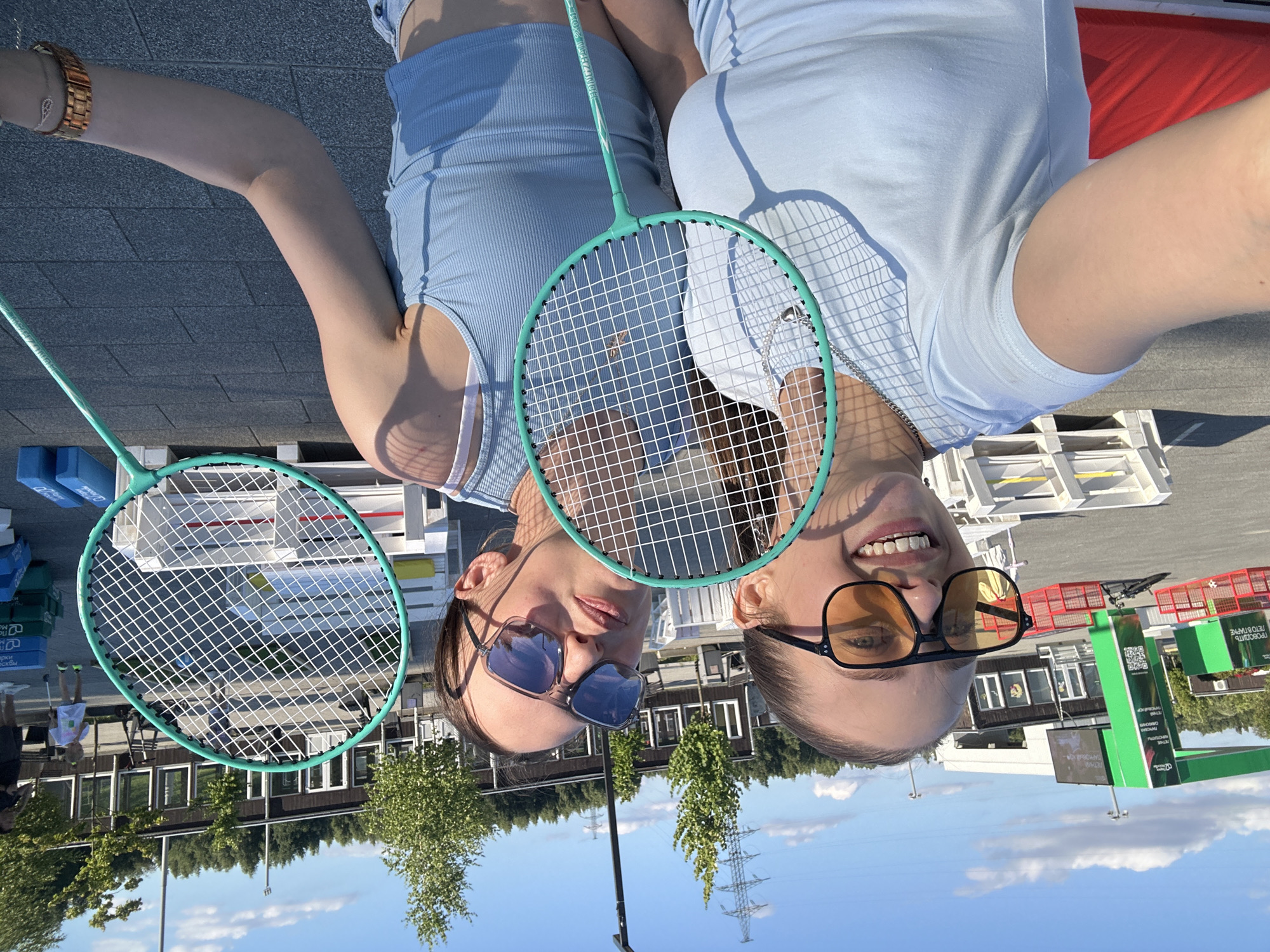 Two young women are posing for a selfie outdoors, holding badminton rackets. The image is taken from a low angle, with the women's heads in the foreground and their bodies extending away. Both women are smiling and appear to be enjoying themselves. They are both wearing light blue athletic tops. The woman on the left is wearing sunglasses. The woman on the right is also wearing sunglasses.
The background shows a paved area with some wooden pallets and what appear to be blue blocks. Beyond that, there is a building with windows, and behind the building, there are trees and a clear blue sky with some clouds. A power line tower is visible in the distance. The sun is shining brightly, suggesting it is daytime.
The overall scene appears to be a recreational area, possibly a sports complex or an outdoor event. The presence of badminton rackets suggests they might be about to play or have just finished playing the sport. The setting seems to be casual and summery.
Some text is visible on green signs in the background, which are advertising something related to a "summer festival" or "summer games" in Russian. One sign includes a QR code. The location context is Khimki, Russia.