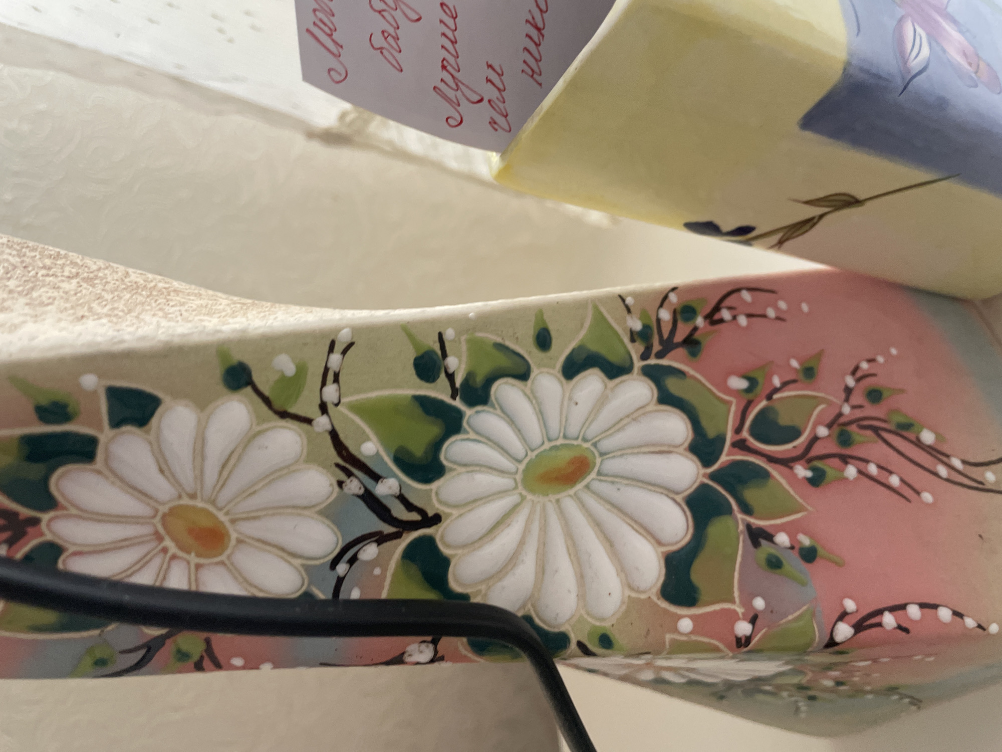 The image shows a close-up of two decorative vases. The lower vase has a floral pattern of white daisies with green leaves and pink background, while the upper vase has a yellow and blue floral design. A piece of paper with red text is partially visible above the upper vase. The scene is indoors, possibly in a residential setting, but the location within Olginka, Russia, cannot be determined from the image alone.