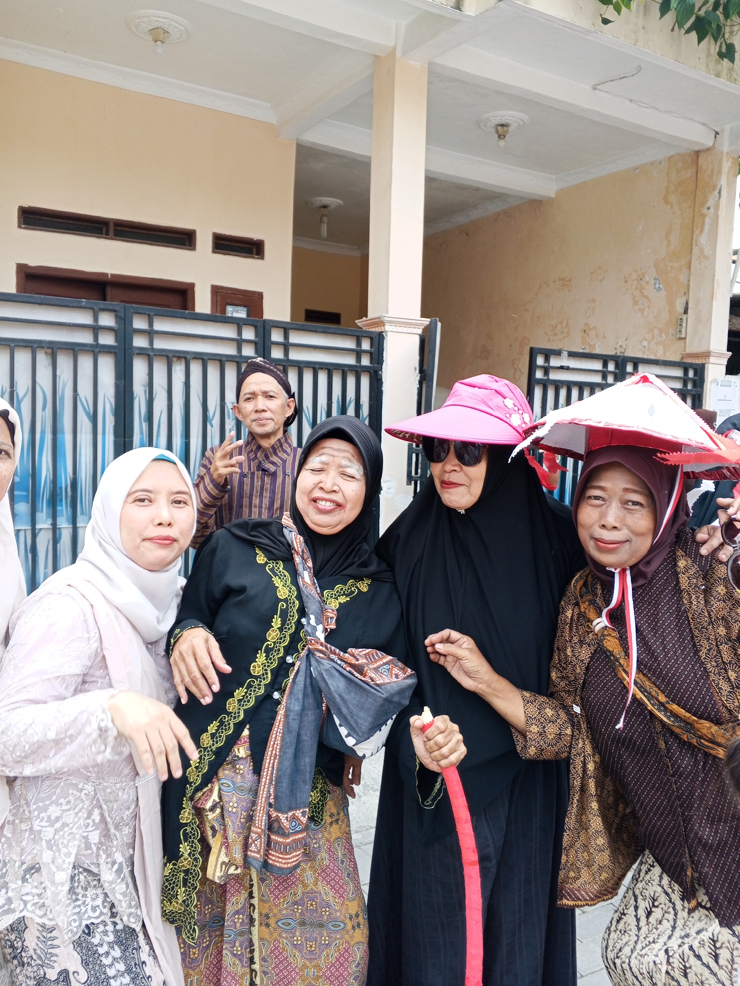 Five individuals are pictured outdoors in front of a building with a cream-colored facade and a white awning. On the left, two women are visible. The woman in the foreground wears a light pink lace top and a matching headscarf. To her right, another woman, also wearing a white headscarf, smiles directly at the camera. Behind them, a man wearing a traditional patterned brown and purple striped shirt and a dark head covering is gesturing with his right hand. To the right of the man, a woman in a black hijab smiles with her eyes closed, her face powdered to resemble aging. She holds a gray and patterned scarf over her shoulder. Next to her, a woman wearing a brown patterned batik top and a maroon headscarf is smiling. She wears a conical red and white hat adorned with fringe and appears to be holding a long, red object that resembles a stick or staff. Behind the group, a black metal gate is visible, with a portion of a building and greenery in the background. The lighting suggests it is daytime. The overall scene appears to be a gathering or celebration.