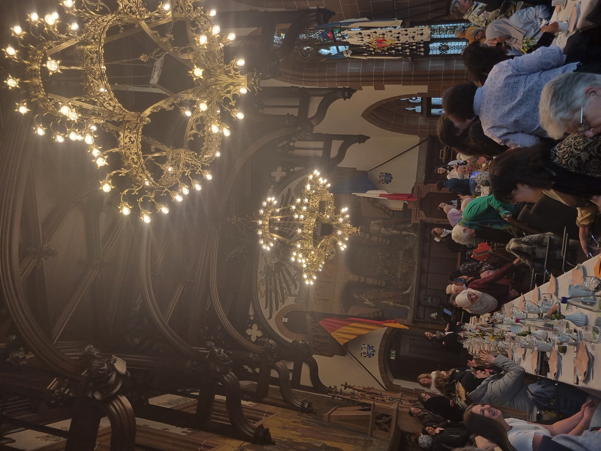 The image shows a banquet hall interior, likely the Saarbrücken City Hall (Rathaus) in Saarbrücken, Germany, given the architecture and German flags. Two large ornate chandeliers illuminate the space. Tables are set for a meal, and people are seated and dining. An elaborate mural adorns the wall behind the tables.