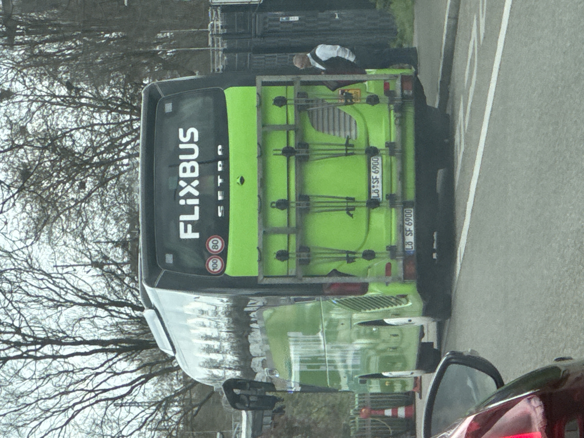 A lime green Flixbus is stationary on a road in Kehl, Germany. The bus has a luggage rack on the rear. A man wearing a white shirt and dark trousers is standing near the back of the bus. The license plate reads "LÖ SF 6900". Trees with bare branches are visible in the background on the left. The scene appears to be captured from inside another vehicle.