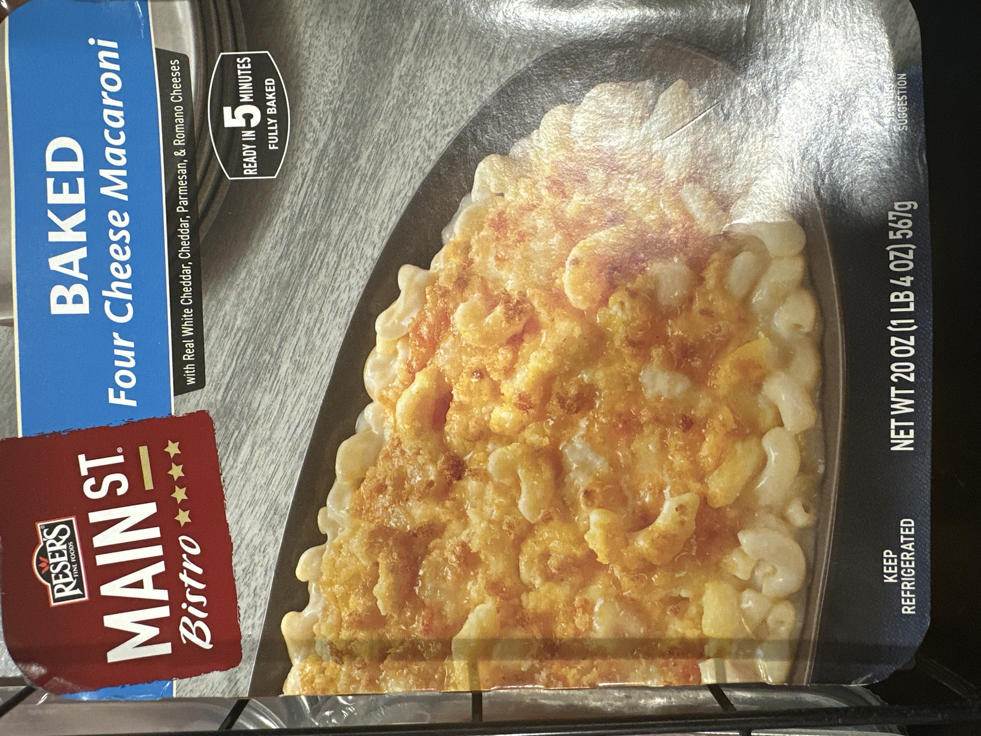 The image is a close-up, eye-level shot of the front of a box of Reser's Main St. Bistro Baked Four Cheese Macaroni. The box is predominantly dark blue with white text, and a large portion of the image is taken up by a photograph of the macaroni and cheese itself. The macaroni and cheese appears to be golden brown on top with visible elbow macaroni and melted cheese. Text on the box includes "BAKED Four Cheese Macaroni," "with Real White Cheddar, Cheddar, Parmesan, & Romano Cheeses," and "READY IN 5 MINUTES FULLY BAKED." There is also a logo for Reser's Fine Foods and "MAIN ST. Bistro" with stars. The bottom of the box has text reading "KEEP REFRIGERATED," "NET WT 20 OZ (1 LB 4 OZ) 567g," and "SERVING SUGGESTION." The background is a gray, wood-grain pattern. The lighting is even, and the image is sharp and clear. The location of Camp Hill, United States of America is not depicted in this image, as it shows only the product packaging.