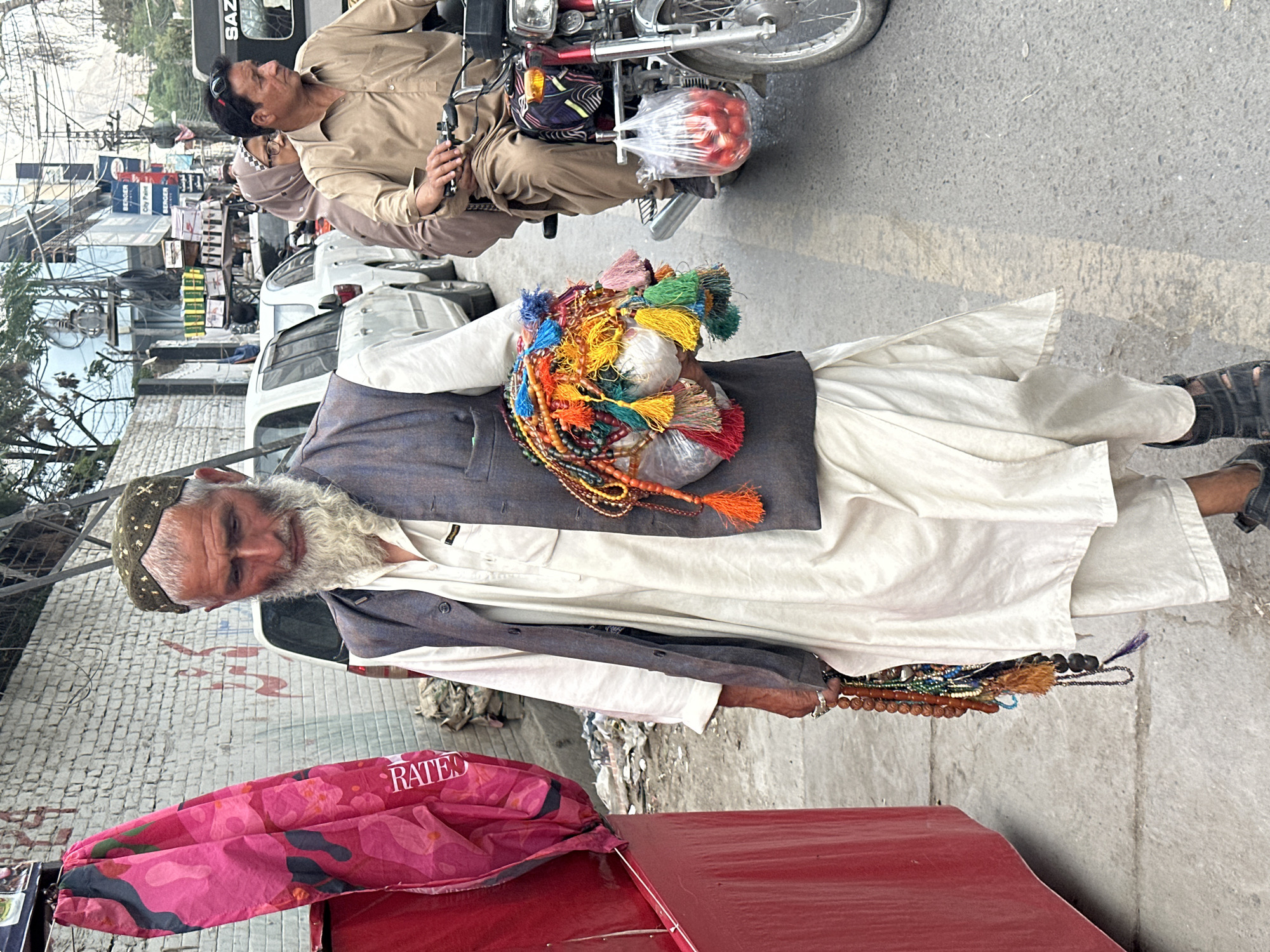 In Quetta City Tehsil, Pakistan, an elderly man with a gray beard stands near a red surface, possibly a cart. He is wearing a cap, a light-colored shirt, a gray vest, and long white pants. He holds strands of colorful prayer beads or rosaries. More prayer beads with tassels are bundled on his chest. A motorcycle with two riders and additional vehicles are visible in the background. The surroundings are urban with buildings and utility lines.