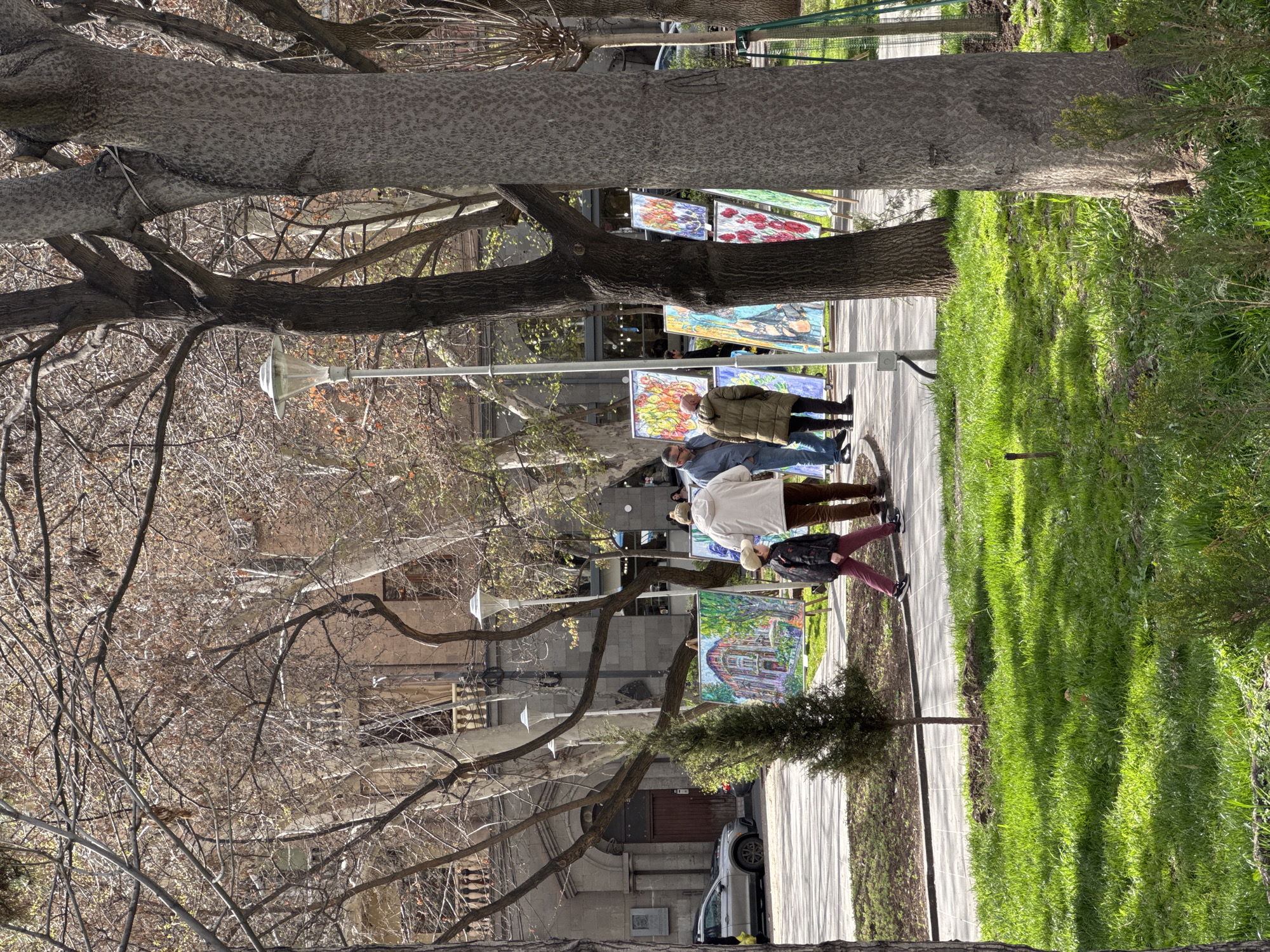 Captured from a ground-level perspective, the scene unfolds along a pedestrian walkway flanked by trees and buildings. Several individuals are observed viewing a display of artwork exhibited along the walkway. The artwork consists of paintings arranged on stands.
Amidst the display, there is a man wearing a mask, indicating potential health concerns or regulations. A car parked near the edge of the walkway suggests the possibility of street parking in the vicinity.
The presence of deciduous trees with bare branches implies the season is either late fall or early spring. Lush green grass on one side of the walkway indicates a managed green space, possibly a park or garden area. The buildings on the opposite side of the walkway suggest an urban environment.
The location is Yerevan, Armenia.