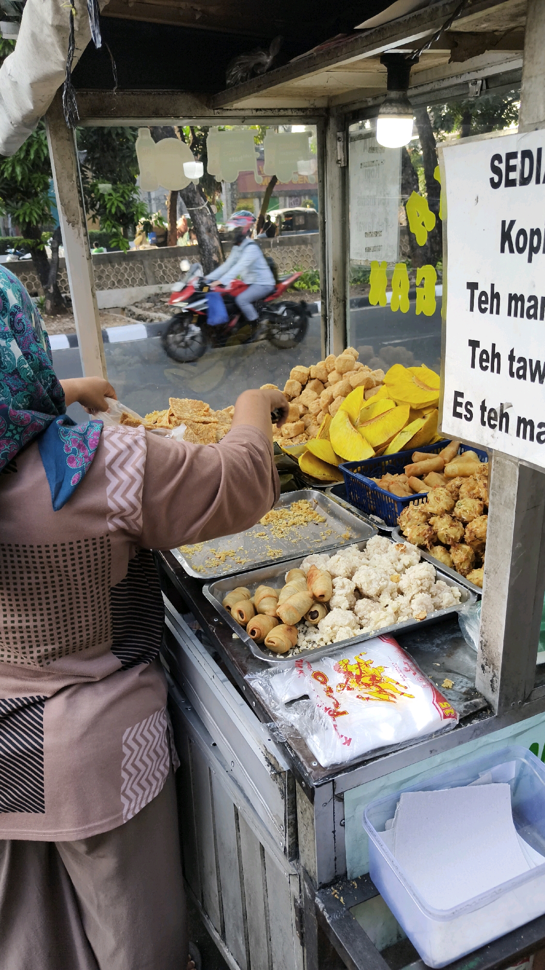 A street food cart is displayed in South Jakarta, Indonesia. A vendor, wearing a hijab and a patterned long-sleeved shirt, is arranging food on a tray. The cart is filled with various fried snacks, including what appears to be tempeh, breadfruit, and other fried items. Behind the food, a motorcycle rider is visible through the glass enclosure of the cart, moving along a street lined with trees and a concrete barrier. A sign on the cart lists "SEDIA KOPI", "Teh man", and "Es teh man". In the foreground, a plastic bag with a yellow graphic is placed on the counter.