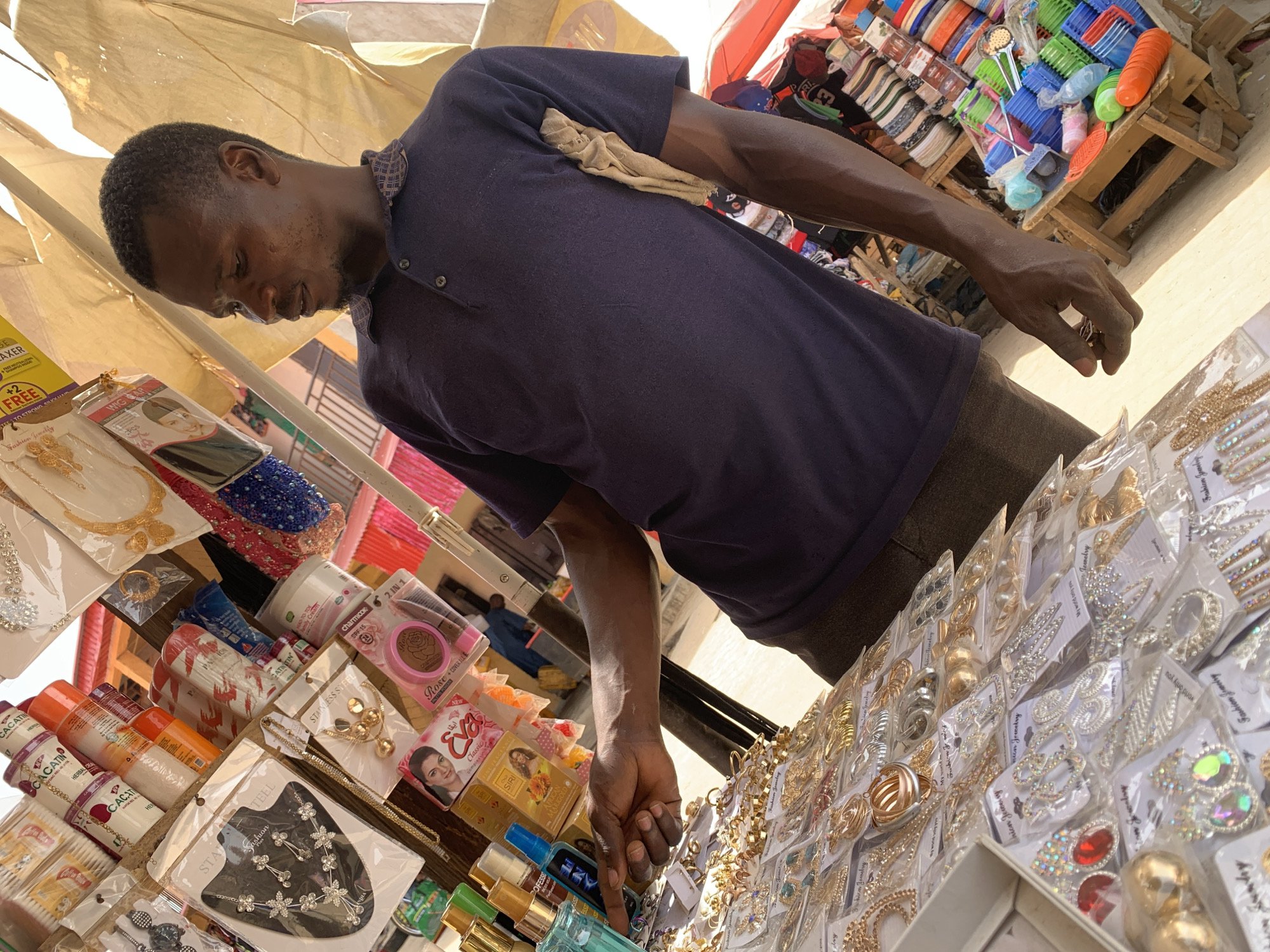 The image shows a man at an outdoor market stall in Kafanchan B, Nigeria, surrounded by an array of goods. The man, with short dark hair and a slight beard, wears a dark blue polo shirt with a patterned collar and dark trousers. A light-colored cloth is tucked into his shirt. He is leaning over the stall, intently looking down and reaching his right hand towards items on display, suggesting he is a vendor attending to his wares or arranging them.
The stall is densely packed with merchandise, predominantly fashion jewelry, cosmetics, and personal care products. On the left side, various creams and lotions are visible, with brands like "CACATIN," "HERBALDE," "charmlook 2 IN 1," "Rose," "Eva Classic," and "SIRI AFRICAN MARIGOLD" identifiable. Some products mention "SPF 15." A hair relaxer or similar product with text "AXER," "FREE +2," and "TO STRONG SHUVUAIN" is also present.
The right side of the stall is filled with numerous pieces of jewelry, including necklaces, earrings, and hair clips, mostly gold or silver-colored, often embellished with stones. Packaging frequently states "Fashion Jewelry" and "STAINLESS STEEL," with phrases like "My World Only You" also visible.
In the background, beyond the man's stall, there are stacks of colorful plastic household items like baskets and bowls, indicating a wider market setting. The scene is illuminated by bright natural light, suggesting it is daytime. The image has a tilted perspective.