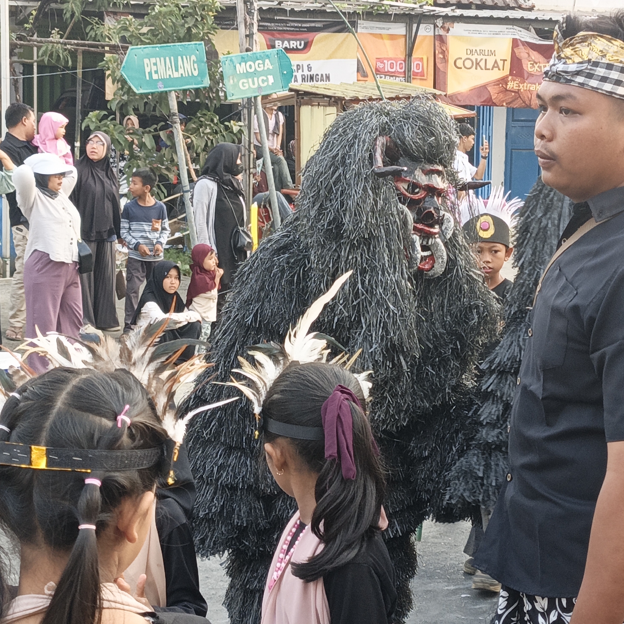 In Randudongkal, Indonesia, a street scene unfolds with a mix of people and a large, dark, costumed figure. The figure, wearing a black, shaggy, full-body costume with a red and white mask, stands prominently in the center. A man in a black shirt and patterned pants stands to the right of the costumed figure, facing partially away from the camera. Several children and adults are visible in the background, some observing the costumed figure. Two young girls in the foreground are also looking towards the figure. Green directional signs are mounted on poles, one reading "PEMALANG" and another "MOGA GUCH". Behind the signs and people, stalls or shops are visible. The environment appears to be an outdoor public space, possibly a street or market.