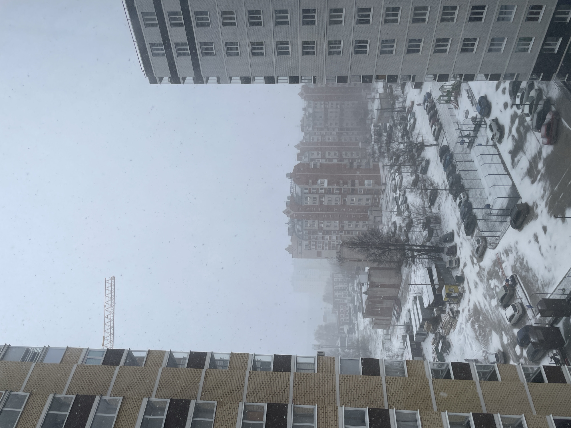 The image captures a winter scene in a city, likely Izhevsk, Russia, as suggested by the architecture. Snow is falling heavily, obscuring the background and creating a white haze. Two buildings frame the view. The top building is a light-colored, multi-story apartment complex with rows of windows. The bottom building, closer to the viewer, features a beige brick facade with black window frames. In the center, obscured by the snow, are other buildings of varying heights and colors, including a red-roofed building. A tall, yellow construction crane is visible in the distance, rising above the lower building. Below and to the right of the crane are cars parked in a snow-covered lot. A playground is visible in the middle ground. The entire scene is blanketed in snow, creating a monochrome, wintery landscape.