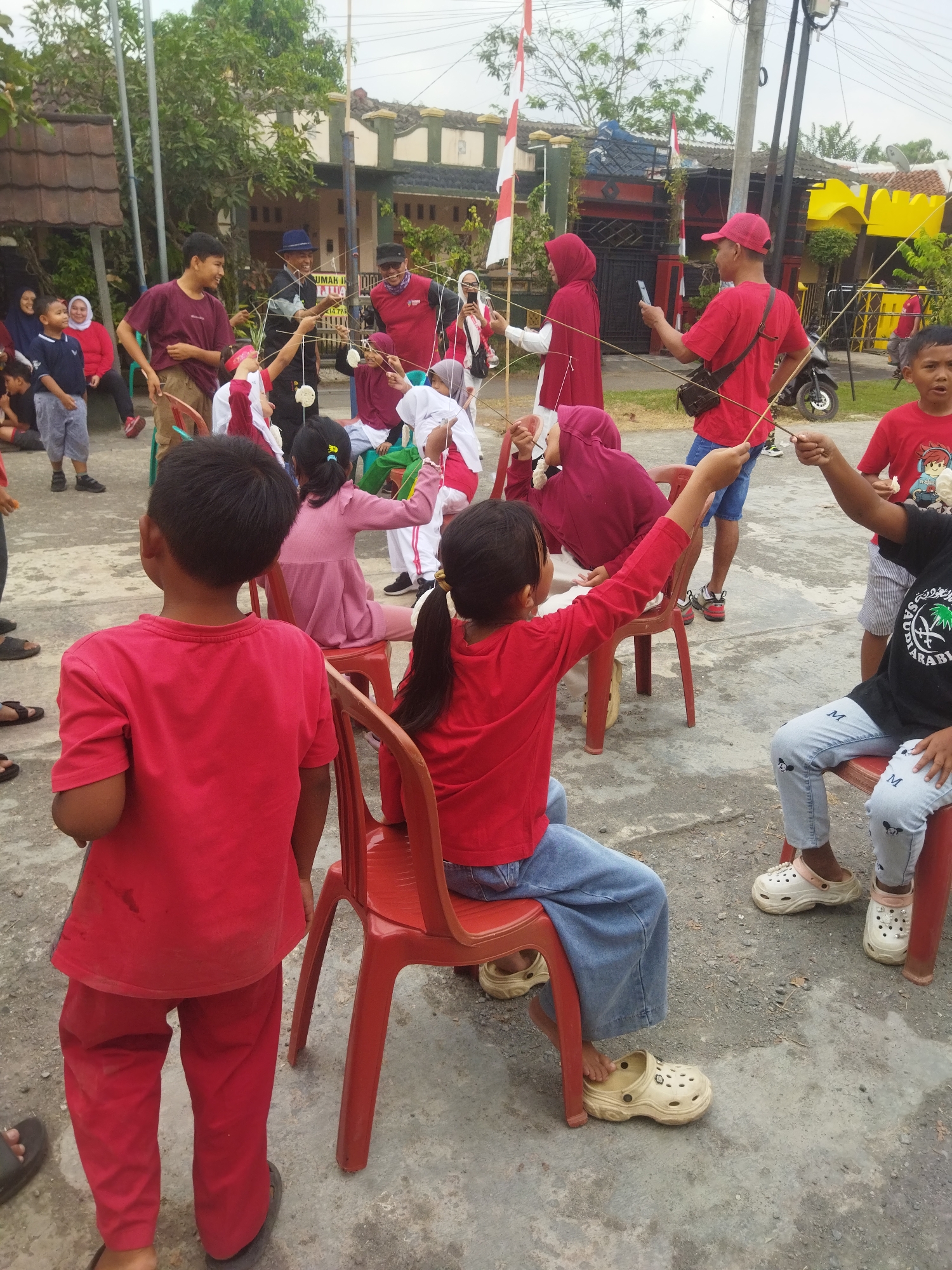 This is a photograph taken at a children's game event in Banjar, Indonesia. The scene appears to be an outdoor gathering, possibly a community event or celebration, with several children and adults participating in a game. The game involves using sticks to hold small, white food items (possibly crackers or marshmallows) near the mouths of participants.
In the foreground, a young boy in a red t-shirt and red pants is facing away from the camera, with his back to the viewer. To his right, a young girl also in a red shirt and blue jeans is seated on a red plastic chair, actively engaged in the game with a stick in her hand. Another child, in a black t-shirt and light blue jeans, is seated to the right of the girl, also participating.
In the middle ground, more children and adults are visible. A girl in a pink dress is seated on a red chair, reaching out with a stick. Several adults are observing or supervising the game, some of them holding their phones. One man in a red t-shirt and a red cap is holding a stick and looking at his phone. Another man in a blue shirt and hat is also participating in the game. A woman in a red hijab is also present.
The background shows a residential area with houses, some decorated with Indonesian flags, suggesting a festive occasion. There are trees and utility poles visible, indicating an urban or suburban setting. The ground is paved with concrete. The overall atmosphere appears to be one of fun and community engagement.