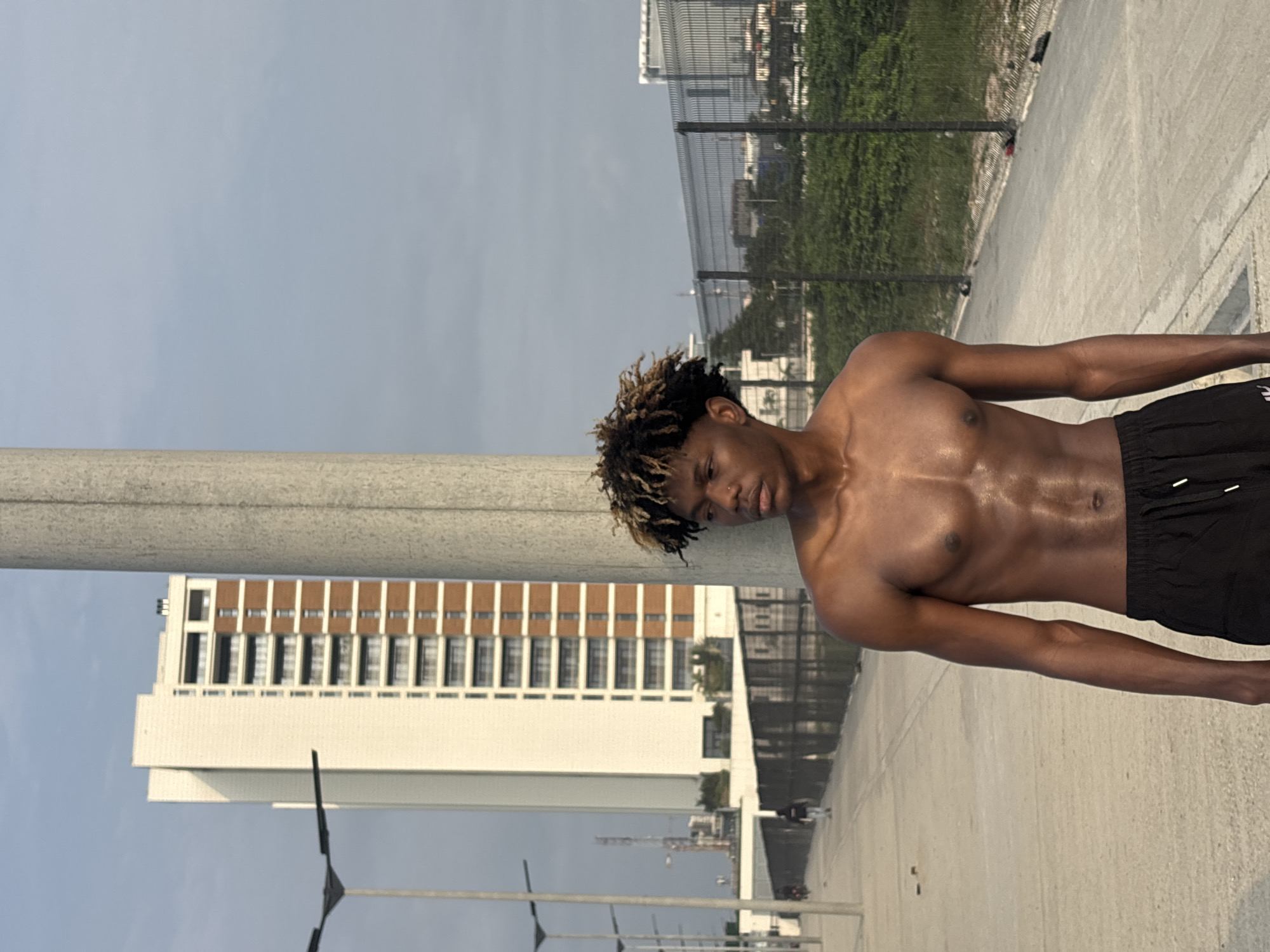 The image depicts a young Black male, shirtless and wearing black athletic shorts, standing next to a concrete utility pole. His body is muscular, with visible abdominal definition and sweat. His hair is styled with dreadlocks, some of which are lighter in color. He is looking to his right, with a neutral expression.
In the background, a tall, modern white and brown building with numerous balconies stands prominently. To the left of the building, a street lamp with multiple lights is visible. A paved walkway stretches into the distance, with several figures walking along it, including one in the mid-ground wearing dark clothing. A dark metal fence runs along the right side of the walkway, separating it from an area with dense green foliage. Beyond the fence and foliage, other buildings and structures are visible, suggesting an urban environment. The sky is overcast with a pale blue and grey hue. The scene appears to be an outdoor public space in the city of Alaguntan, Nigeria.