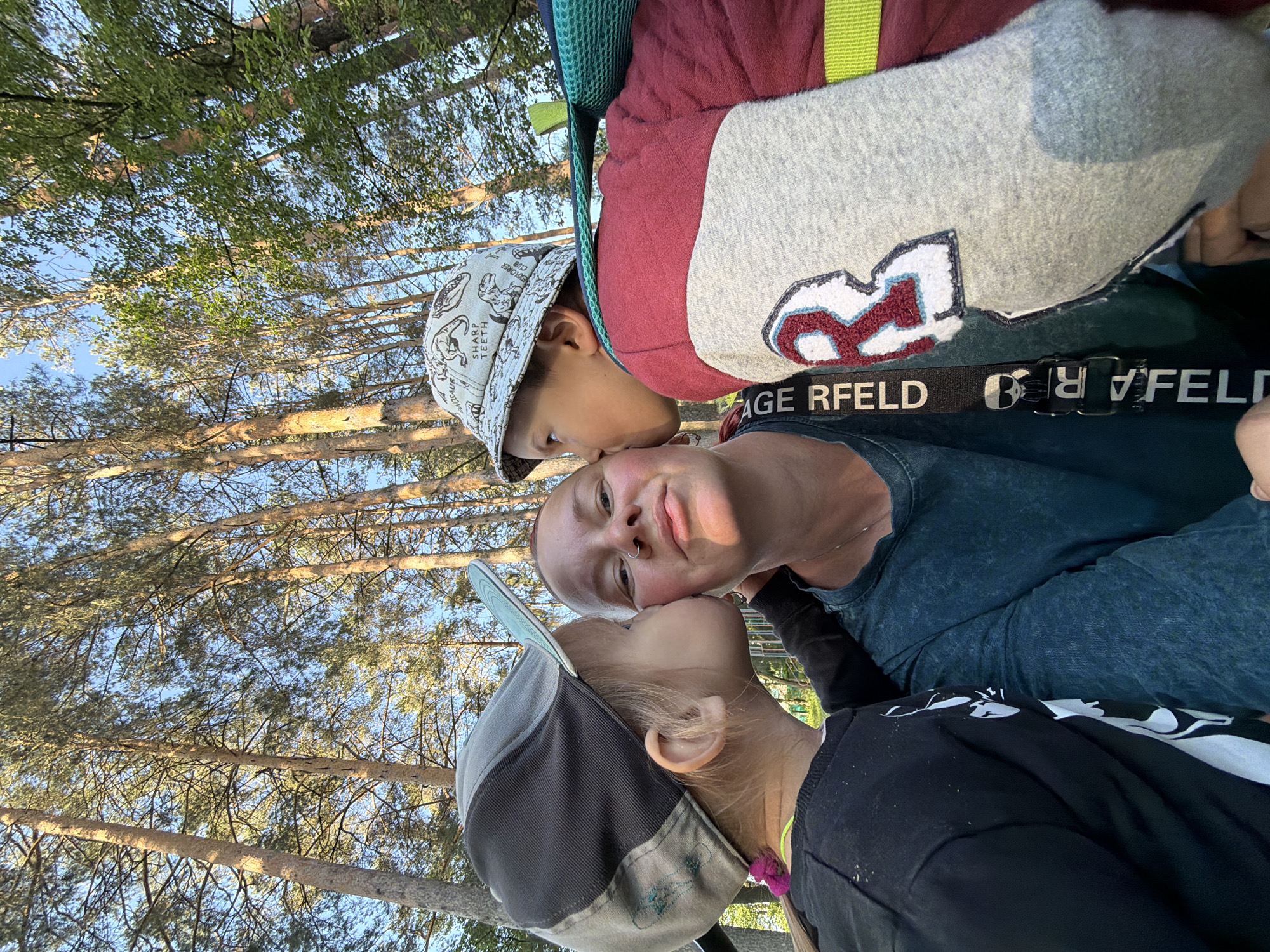 A woman is photographed from a low angle as two children kiss her face. The background shows tall pine trees against a blue sky. The older child on the top left is wearing a dinosaur-patterned bucket hat. The woman wears a blue-greenish top. The child on the bottom right wears a black and gray baseball cap and a black long-sleeve shirt. Based on the environment, this photo could be taken in a wooded area, potentially within a park or recreational area in Chaikovsky, Russia.