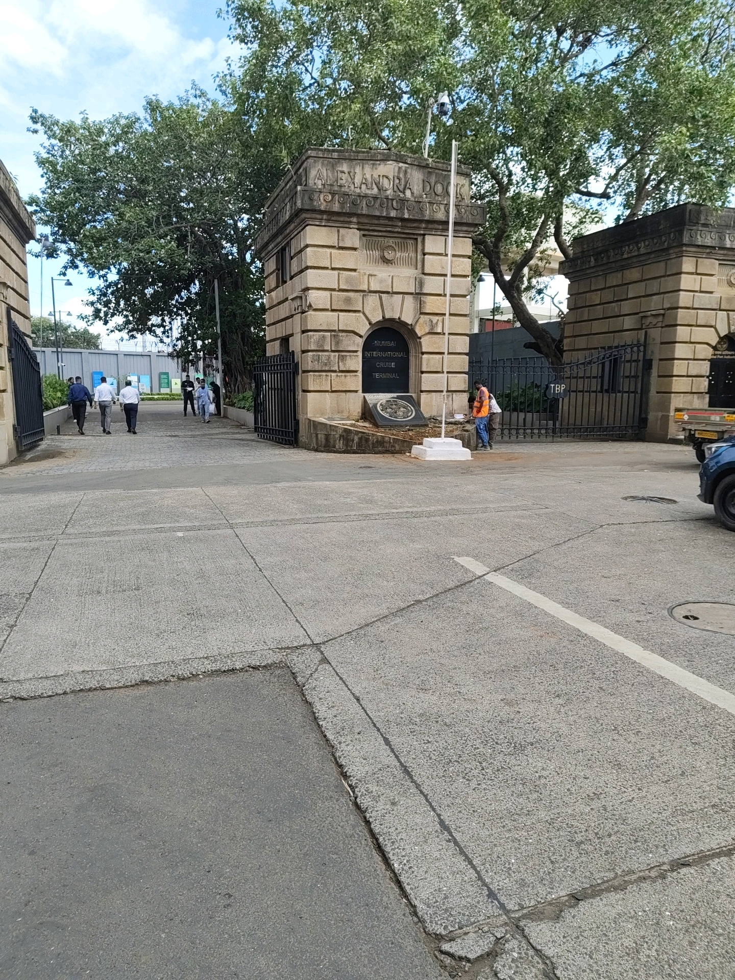 The image shows the entrance to the Mumbai International Cruise Terminal, also known as Alexandra Dock. The entrance is flanked by two large stone pillars, with intricate carvings and inscriptions. A large tree with dense foliage stands behind the pillars, providing shade. Several people are seen entering and exiting the terminal. Some are dressed in formal attire, suggesting they might be travelers. Others appear to be workers, with one person in an orange vest. A truck is parked to the right of the entrance. The ground is paved with concrete slabs, and a white line marks a lane. The sky is partly cloudy. The overall impression is of a busy port facility on a sunny day.